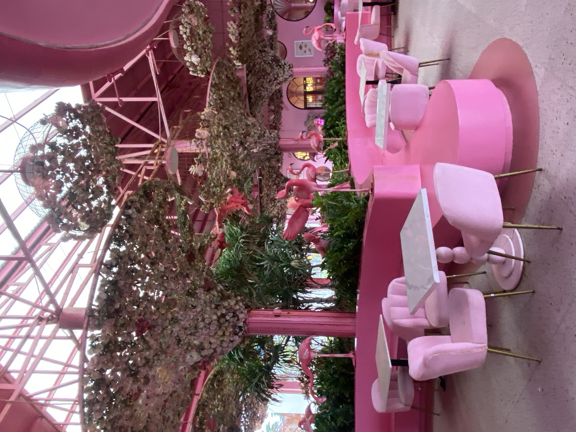 This image captures a vibrant, pink-themed interior, characteristic of a whimsical cafe or restaurant in Tien Thanh Ward, Vietnam. The entire space is dominated by various shades of pink, from light pastel seating to bright fuchsia structural elements and decor.
The area features a high ceiling with visible pink arched beams and a complex truss structure, suggesting a spacious, possibly atrium-like design that allows for significant natural light, indicating daytime. Abundant artificial green foliage, vines, and white flowers are draped and hung from the ceiling and structures, creating an immersive, garden-like canopy. Decorative birdcages, some appearing to contain fans or additional greenery, are also suspended overhead.
The seating arrangements consist of numerous pink chairs with gold or black legs, paired with small white tables. Some chairs are plush and upholstered, while others have a more modern, streamlined design. Additionally, there are larger, rounded pink booth-style seating areas integrated into the space. Throughout the cafe, particularly within planters and amidst the greenery, several pink flamingo statues are prominently displayed, reinforcing a tropical and playful theme. Pink-painted pillars and dividers further section the area.
The overall atmosphere is cheerful, unique, and highly stylized, designed for dining or relaxation in a distinctly themed environment. No people are visible in the scene, and there is no discernible text.