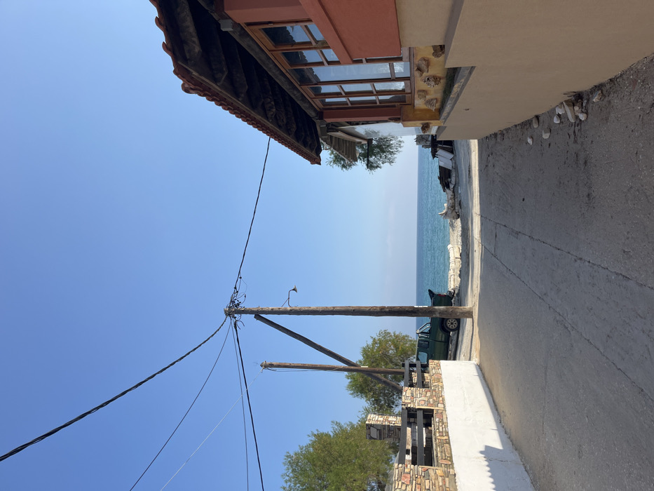 The image shows a narrow street in Limenaria, Greece, with a view of the Aegean Sea in the distance. The street is paved with concrete and lined with low stone walls on either side. A wooden utility pole stands on the left side of the street, with several electrical wires running overhead. A green pickup truck is parked on the left side of the street, facing the sea. The street leads towards the sea, which is calm and blue under a clear, blue sky. The wall on the right side of the street belongs to a building with a red tile roof and large windows. The wall is beige and decorated with seashells. There are no people in the image. The environment is peaceful and serene, with the sun shining brightly. The latitude and longitude coordinates are 40.626, 24.57, which are located in Limenaria, Greece.