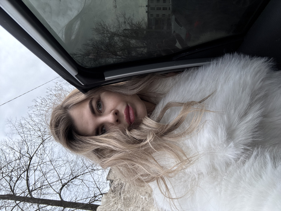 A fair-skinned female with wavy blonde hair and light-colored eyes is depicted in a close-up, frontal view, wearing a white faux-fur coat. She is positioned next to the partially open passenger door of a dark vehicle, looking directly at the camera with a neutral expression. In the upper left background, a large mound of dirty snow and bare deciduous trees are visible against an overcast sky. The tinted window of the vehicle reflects additional bare trees, a multi-story residential building, and other parked cars, including a white vehicle displaying a red taillight. The scene is set in an urban environment in Chaikovsky, Russia, during a cold season.