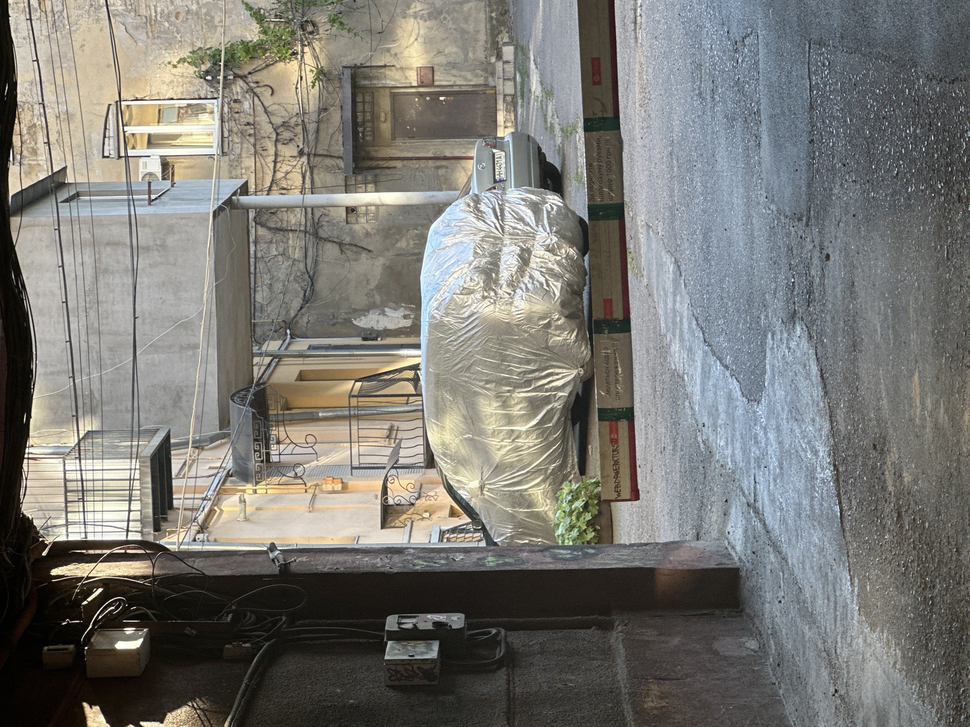 The image captures a narrow courtyard with a rough, grey asphalt ground and textured concrete walls on the right. The walls are adorned with various wires and pipes. In the center of the scene, a car, possibly a silver sedan with license plate "HH 4471A1", is partially covered by a large, crinkled silver tarp. To the left, an apartment building features balconies with wrought-iron railings and windows. Some greenery, including vines climbing the walls and a small bush, adds a touch of nature to the urban environment. In the foreground, electrical equipment, including junction boxes and coiled wires, lies on a ledge. The scene suggests an outdoor, possibly urban, setting, with the presence of buildings, wires, and a car indicating human activity and infrastructure. The lighting suggests it is daytime, with shadows cast by the structures. There is some visible text on a cardboard box leaning against the wall on the right, containing Cyrillic characters that mention "WEBDRIVER", "1000 грн", and "спостереження". The overall impression is one of an old, perhaps slightly neglected, urban courtyard.