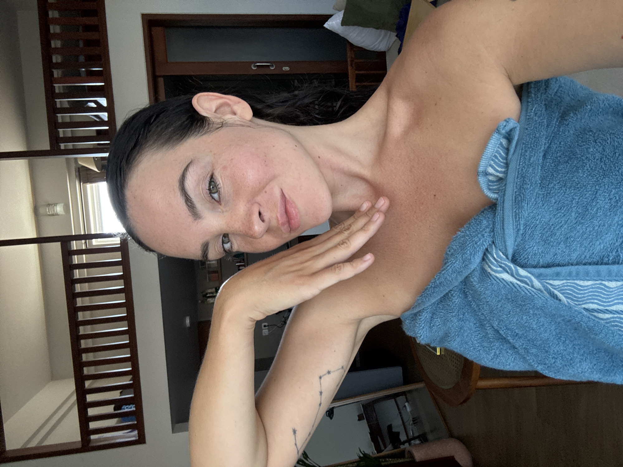 A woman is taking a selfie in a room in North Kuta, Indonesia. She is wrapped in a blue towel and has her left arm raised, with her hand touching her neck. Her hair is dark and wet, and she is wearing light makeup. The background shows a room with wooden railings on a loft and a glass door. There is also a table and chairs in the background. The overall lighting suggests it is daytime.
