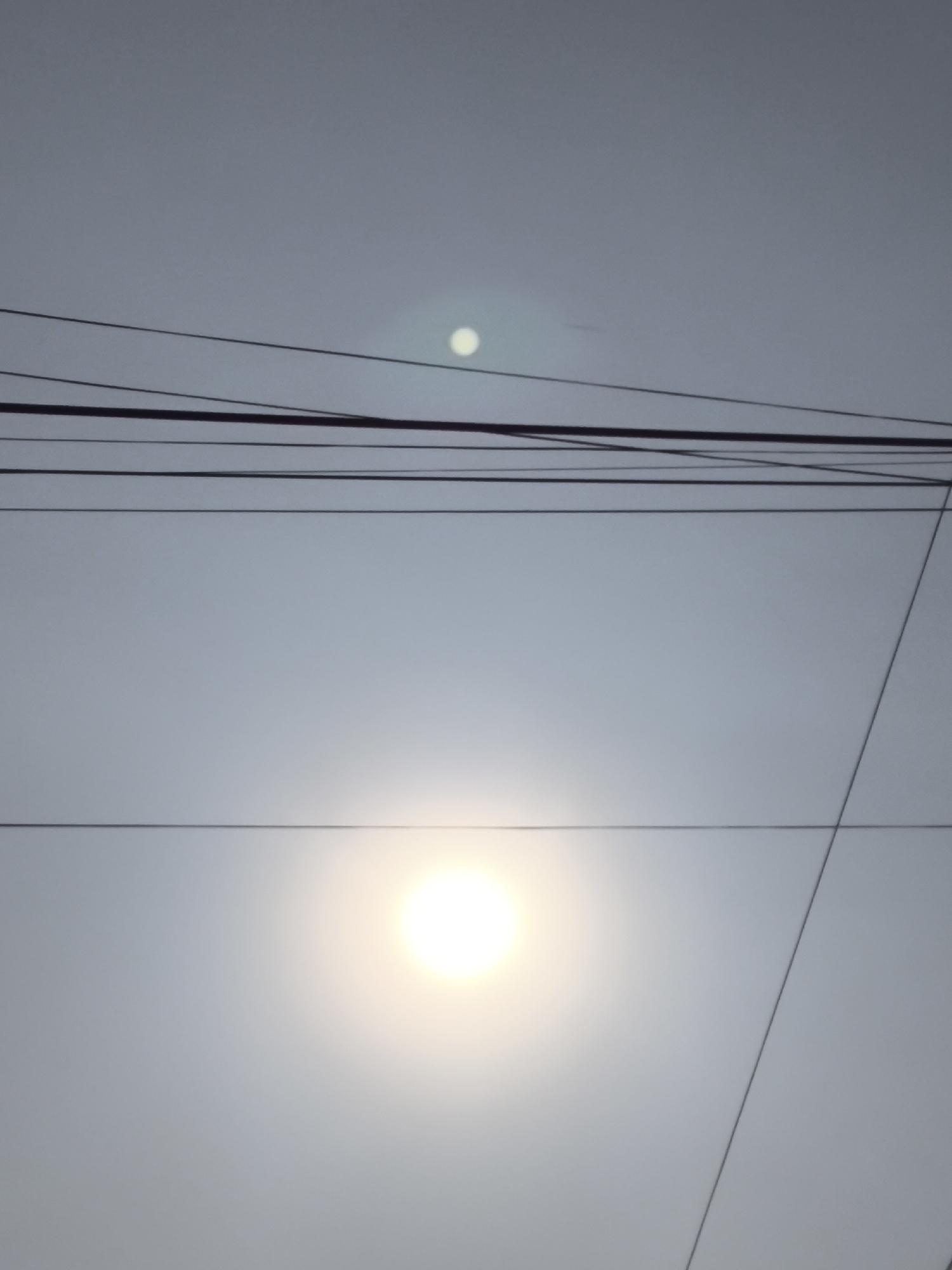 A low-angle shot captures a hazy sky above Cilegon, Indonesia, with the sun as a bright, diffused orb in the lower-middle portion of the frame. Numerous power lines are crisscrossed horizontally and diagonally throughout the sky, some appearing thicker and darker than others. In the upper-middle of the image, a smaller, less distinct circle of light suggests another celestial body, possibly the moon or a lens flare. The overall atmosphere is overcast, with the light suggesting either early morning or late afternoon. No people or specific landmarks are visible in this scene, which focuses on the interplay of the sky, celestial bodies, and the urban infrastructure of power lines.