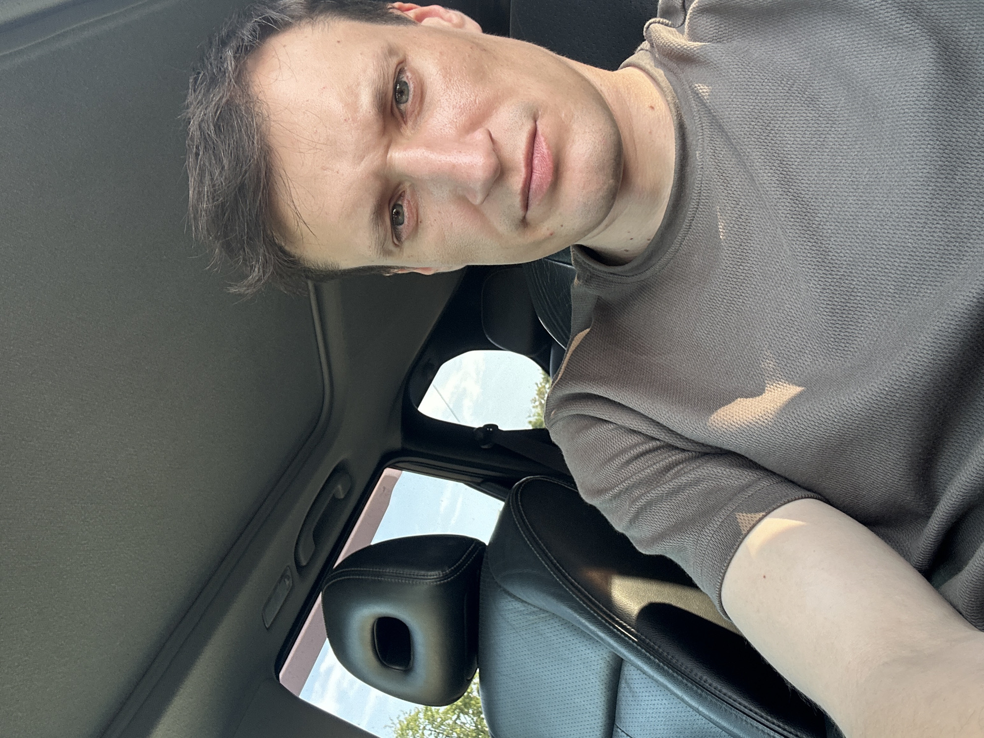 A man with short, brown hair and light green eyes is sitting in the driver's seat of a car. He is wearing a brown, short-sleeved t-shirt. The car's interior is visible, including a black leather seat and headrest with a circular cutout. The sunroof is open, allowing natural light to illuminate the scene. The background shows trees and a blue sky, suggesting an outdoor setting. The location is specified as Shatava, Ukraine.