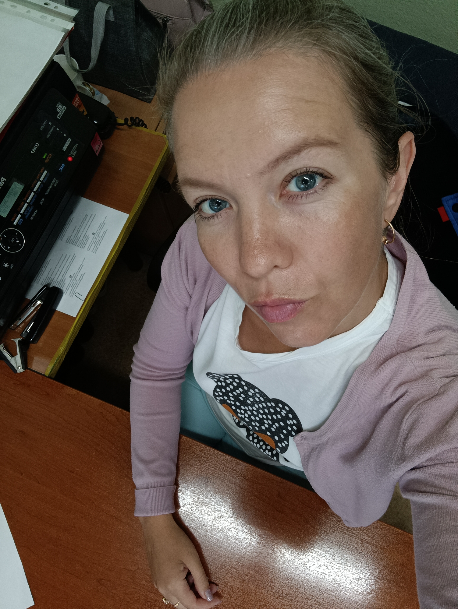 A woman with blonde hair pulled back and blue eyes takes a selfie from a high-angle perspective, sitting at a desk. She is wearing a pink cardigan over a white t-shirt with a black and white owl graphic. On the desk in front of her is a black printer with Cyrillic labels, a stapler, and some papers. A dark grey bag is visible behind the printer. The desk surface is a polished wood. The location is a room in Chaikovsky, Russia.
