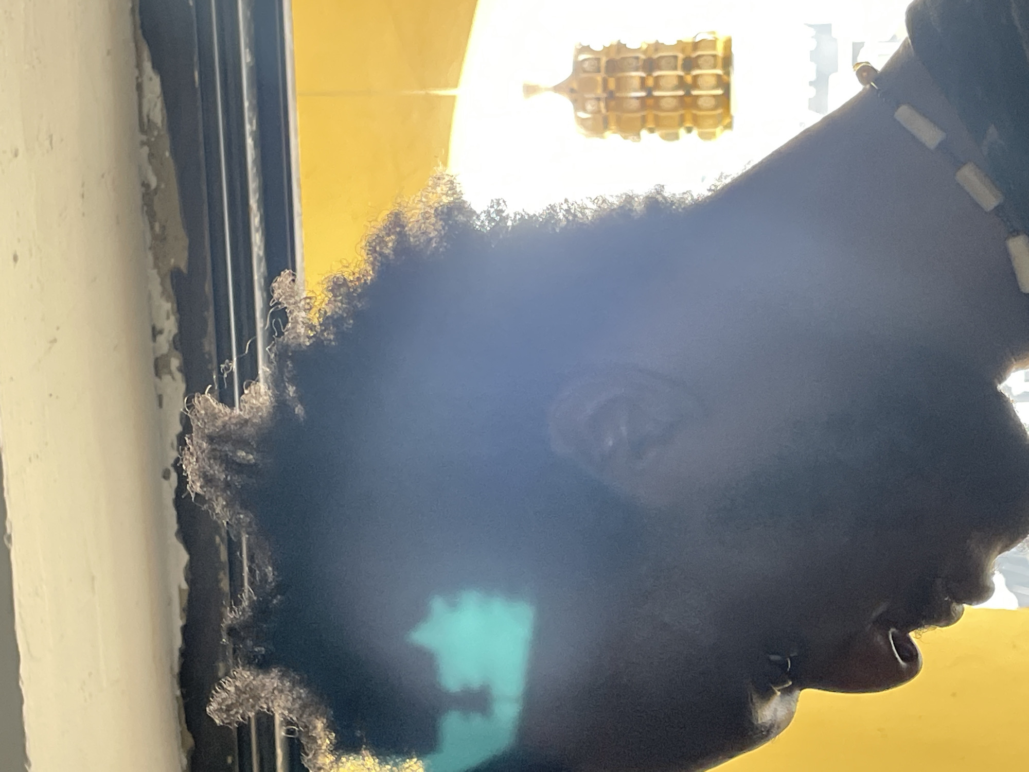 The image is a close-up, profile view of a person with dark, curly hair and dark skin. The person is wearing a necklace with light-colored beads. The background is blurred and consists of a bright yellow surface and a light source, possibly sunlight, with a decorative object hanging in the distance. There is a teal-colored reflection on the person's forehead. The scene appears to be indoors, with a ceiling visible at the top of the image, showing signs of wear and tear. The overall lighting is strong, creating a silhouette effect on the person's face. It is not possible to determine the specific location in Abuja, Nigeria from the image.