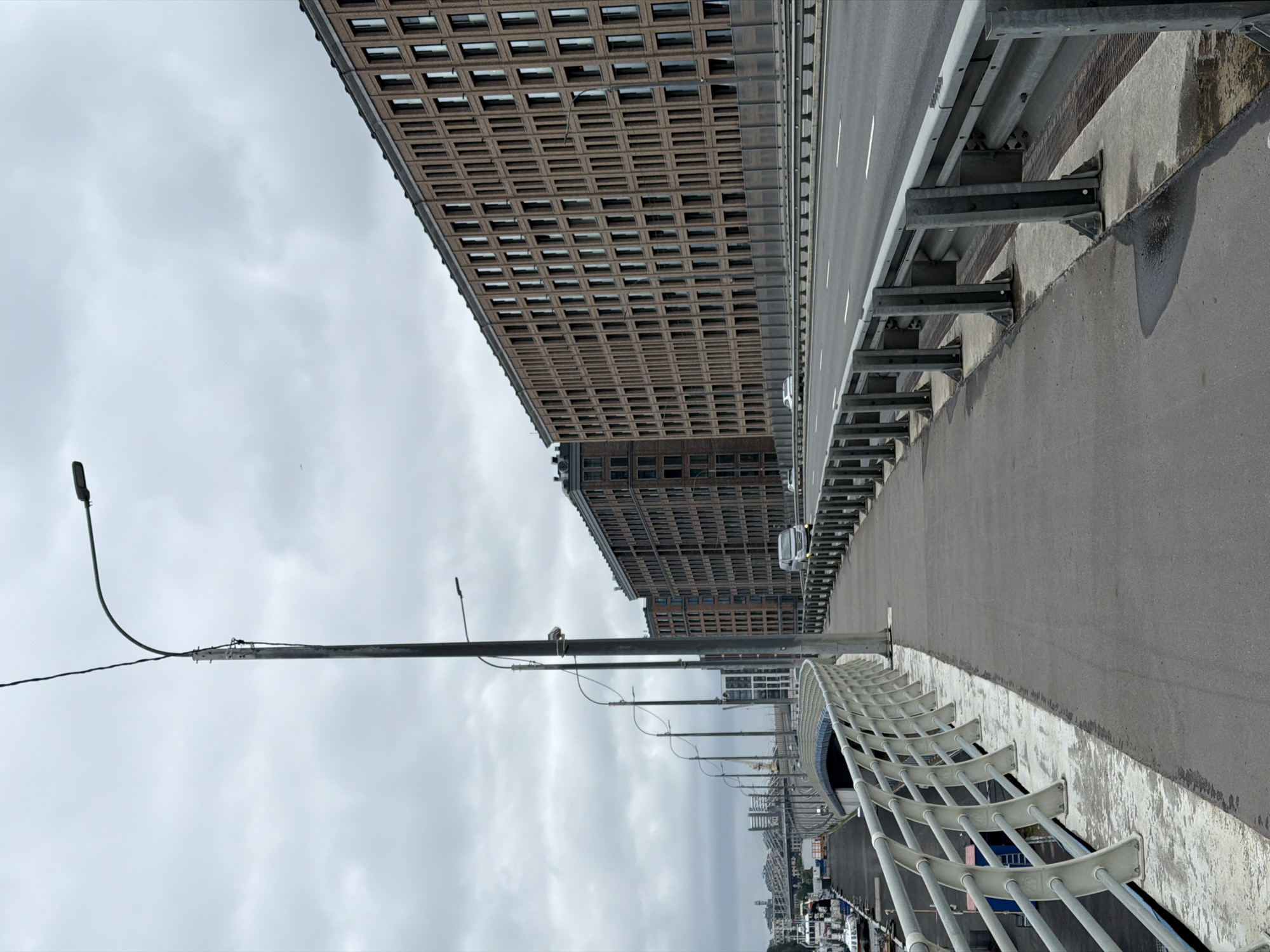 The image depicts a pedestrian walkway running alongside a multi-lane roadway on a bridge in Saint Petersburg, Russia, likely part of the Krestovsky Island area. The walkway has a metal railing along its edge, separating it from the roadway and a lower level which appears to be a harbor or canal with several boats docked. Towering over the roadway and walkway is a modern, multi-story building with a facade of numerous windows. The sky is overcast with heavy clouds, and the lighting is diffused. Several vehicles are visible on the roadway. Multiple lampposts are positioned along the walkway and the road, some with visible wires. In the distance, other bridges and city structures are visible under the cloudy sky.