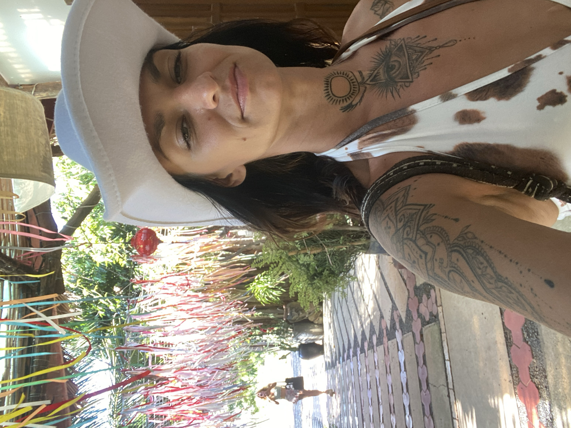 In Mũi Né, Vietnam, a woman with dark hair and wearing a white cowboy hat and a cow-print bikini poses for a selfie. She has tattoos on her chest and arms. In the background, a pathway lined with stone tiles and small red stones leads to a lush, green area with trees and potted plants. Colorful ribbons are strung across an overhead structure, and a red lantern hangs among them. In the distance, another woman walks away from the camera, holding a bag and a phone. The scene appears to be outdoors, possibly in a courtyard or garden area.
