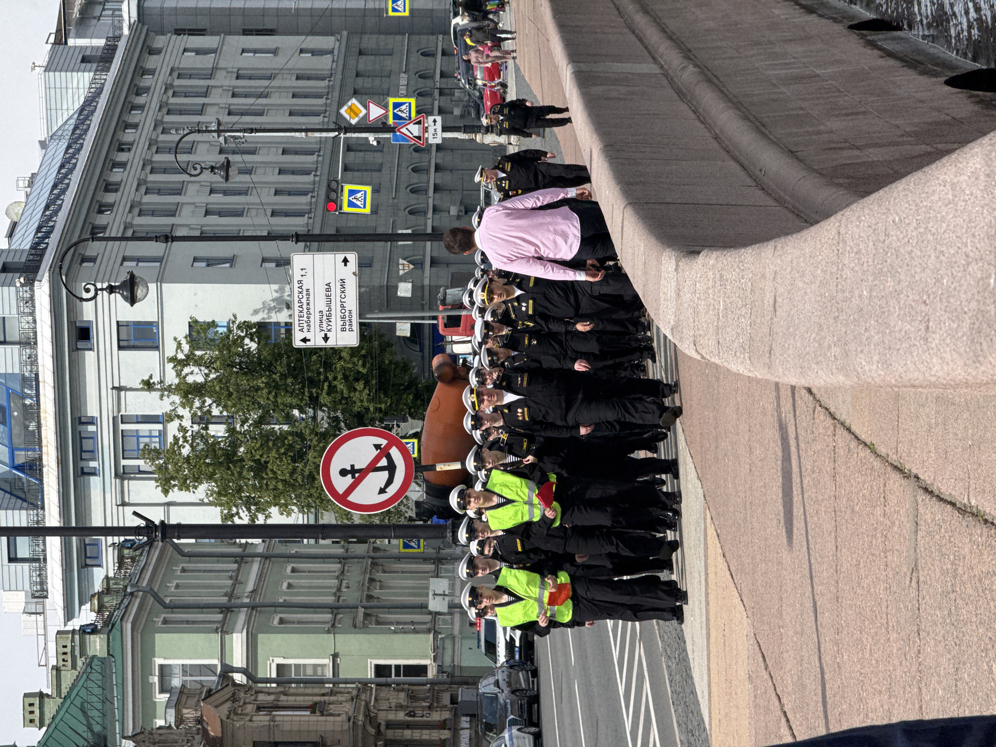 This image depicts an urban scene in Saint Petersburg, Russia, likely along a canal or river embankment, captured from a slightly elevated perspective. The central activity is a procession of a large group of people, primarily men, dressed in dark naval uniforms with white caps and gold insignia. Many of them wear bright yellow high-visibility vests and some carry small red flags, indicating an organized event such as a parade, ceremonial march, or training exercise. A man in a pink striped shirt is seen walking alongside the formation.
The setting features multi-story buildings lining the street, showcasing a mix of architectural styles from classical to more modern. Ornate streetlights punctuate the scene. Visible objects include several road signs: a prominent white directional sign in Russian pointing to "АПТЕКАРСКАЯ 1,1 набережная" (APTEKARSKAYA 1,1 embankment), "УЛИЦА КУЙБЫШЕВА" (KUYBYSHEV STREET), and "ВЫБОРГСКИЙ район" (VYBORGSKY district). Another notable sign is a red circle with a white background and a black anchor crossed out, signifying "No Anchoring." Other traffic signs include a red traffic light, pedestrian crossing symbols, priority road markers, and a steep descent warning.
The time of day is clearly daylight, with bright, even lighting suggesting fair weather. The atmosphere appears orderly and focused, consistent with a formal procession in a public urban space. The Russian text on the signs serves as a strong location cue, confirming the image's context in Saint Petersburg.