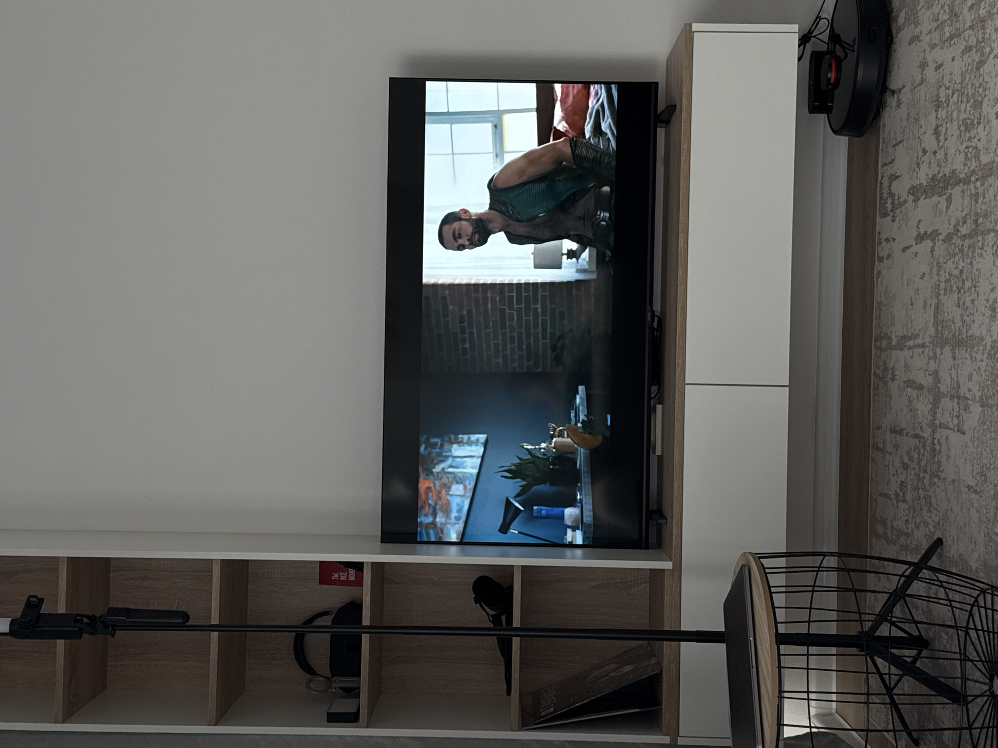This is a medium-shot, eye-level interior photograph of a living room. The scene is dominated by a large flat-screen television displaying a man in a green vest. To the left of the television is a tall, narrow shelving unit with several shelves. On the shelves are various items, including headphones, a red book, a plant, and what appears to be a small lamp. In front of the shelving unit is a black tripod stand with a round wooden table on top, on which a laptop is placed. A small, round, black robot vacuum cleaner is positioned on the floor to the right of the television. The room is furnished with a patterned rug on the floor. The overall lighting is dim, creating a moody atmosphere. The location of this scene is not determinable from the visual information provided.