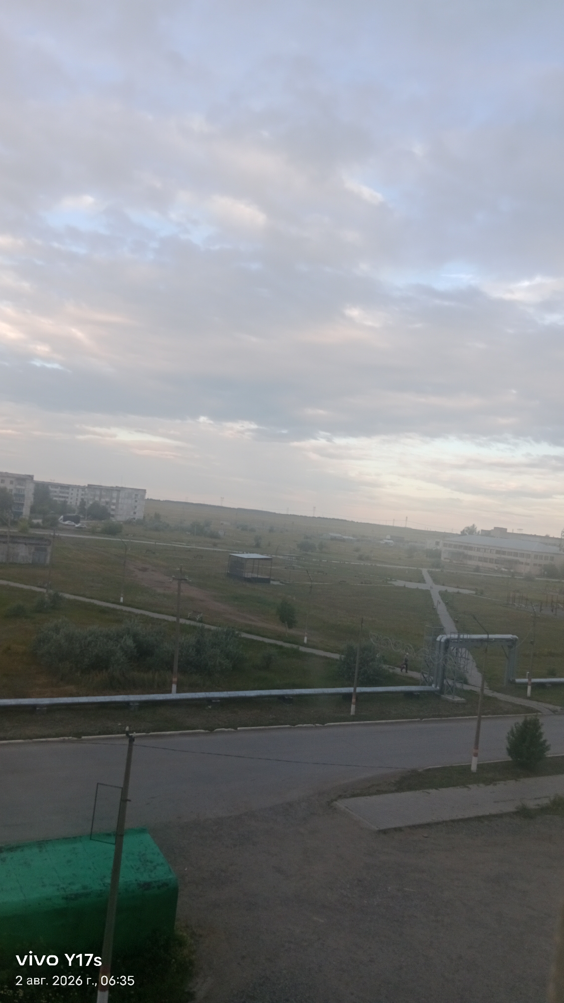 The image captures an outdoor scene in Rudny, Kazakhstan, likely in the early morning given the soft, diffused light and the timestamp on the image. The foreground features a grey asphalt road. To the left, a large, green, rectangular structure is partially visible, appearing to be a storage container or bin. A thin, wooden pole with red and white markings stands next to it, and another similar pole is visible further down the road.
In the middle ground, a paved pathway runs parallel to the road. Beyond the pathway is a wide expanse of green, grassy terrain dotted with low-lying bushes and scattered trees. Several industrial-looking structures are present in the middle ground, including a small, open-sided shelter with a dark roof. Further in the distance, a long, gray building complex or series of apartment buildings is situated on the left side of the frame. On the right, a large, complex industrial structure with metal pipes and supports is visible, with a single person walking nearby. The background consists of rolling hills and an open sky filled with light gray and white clouds, indicating an overcast or dawn sky. The overall impression is that of a semi-industrial or residential area on the outskirts of a city.