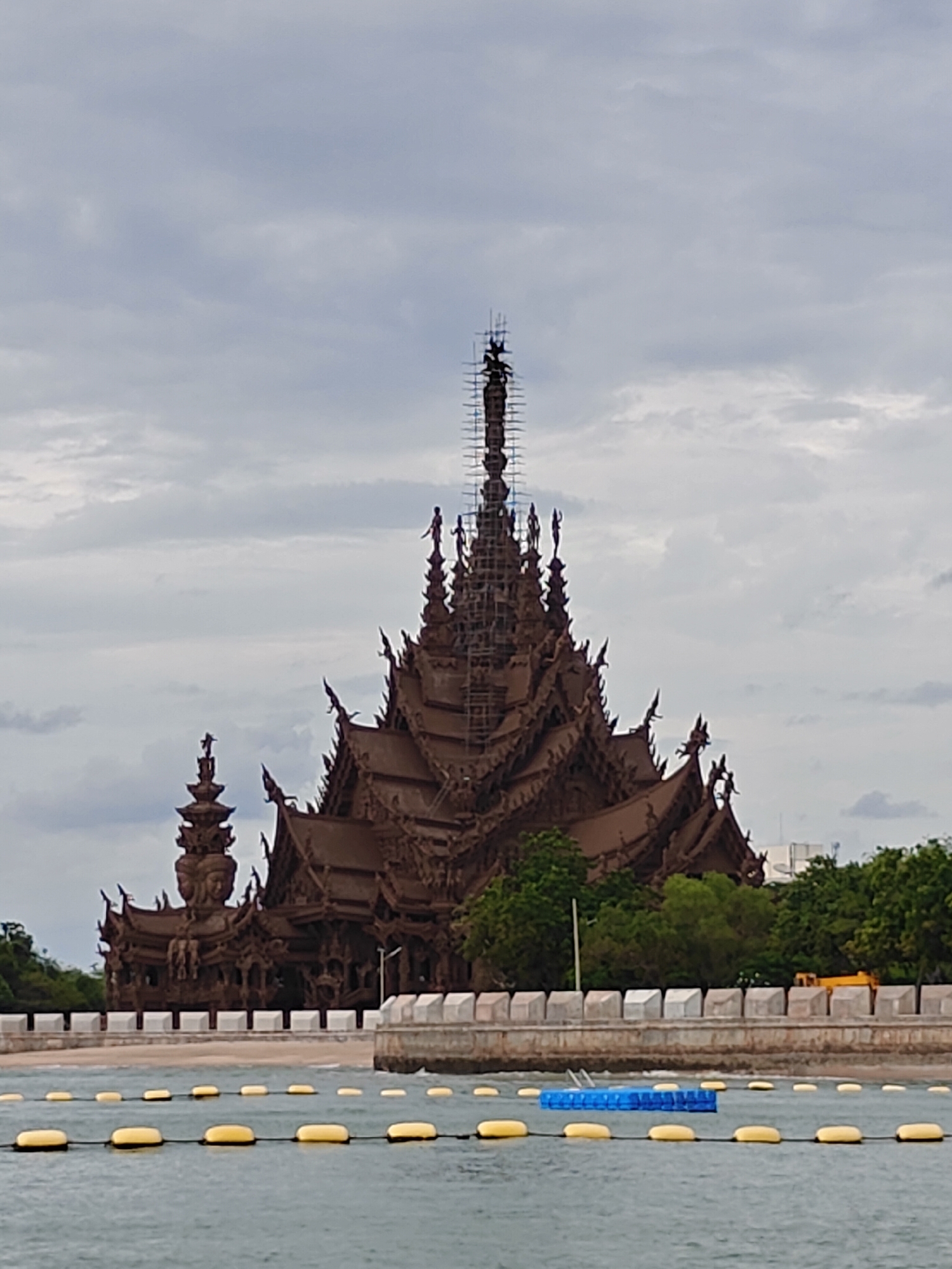 The image shows the Sanctuary of Truth, a colossal wooden temple complex located in Pattaya City, Thailand. The temple, constructed entirely from wood without any metal fasteners, is intricately carved with religious and mythological motifs. Its multi-tiered roof structure features elaborate spires and decorative elements, with scaffolding visible on the tallest spire, indicating ongoing construction or maintenance. The temple is situated on the coast, with a body of water in the foreground marked by yellow buoys and a blue floating platform. Behind the temple, a wall with square white decorative elements runs along the coastline, with lush green trees and some buildings visible in the background. The sky is overcast with clouds, suggesting a dull or partly cloudy day. There are no people clearly visible in the image.