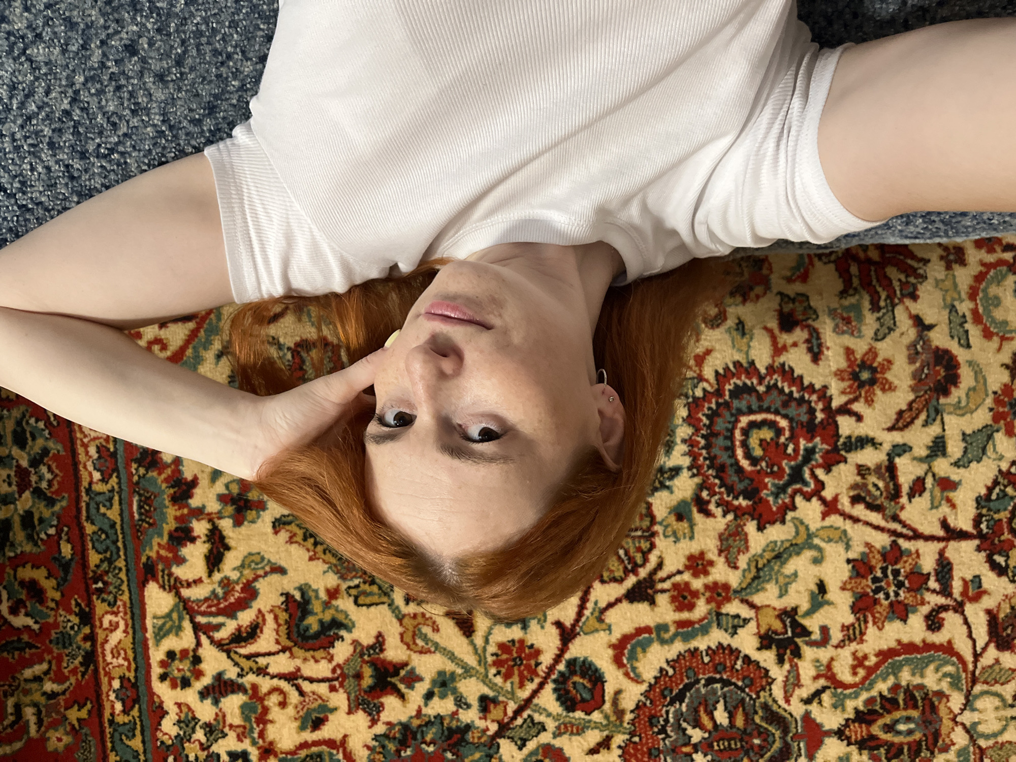 A woman with red hair lies on a sofa in front of a patterned rug. She is wearing a white t-shirt. Her right arm is bent behind her head. The image does not provide any information about the city of Chaikovsky, Russia.