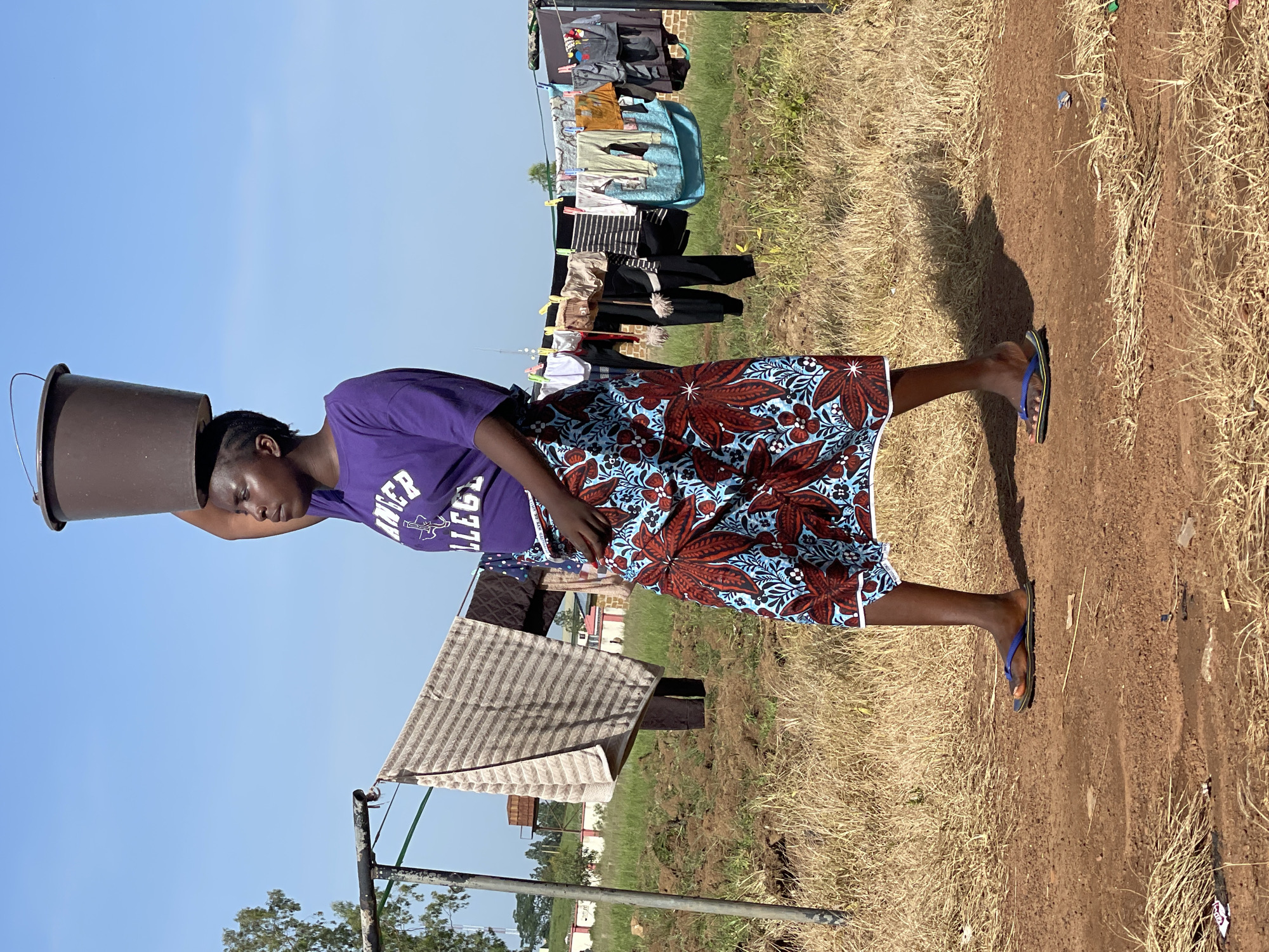 A young woman walks from left to right, carrying a dark brown bucket on her head. She is wearing a purple t-shirt with white lettering that reads "RANCER COLLEGE" and a long, brightly colored skirt with a red and blue floral pattern. She is barefoot in the front, and wearing a blue flip-flop on her right foot. The woman is walking on a dirt path in what appears to be an outdoor area. In the background, there is a clothesline with several items of clothing hanging on it. The sky is clear and blue. This image was captured in the city of Durumi, Nigeria.