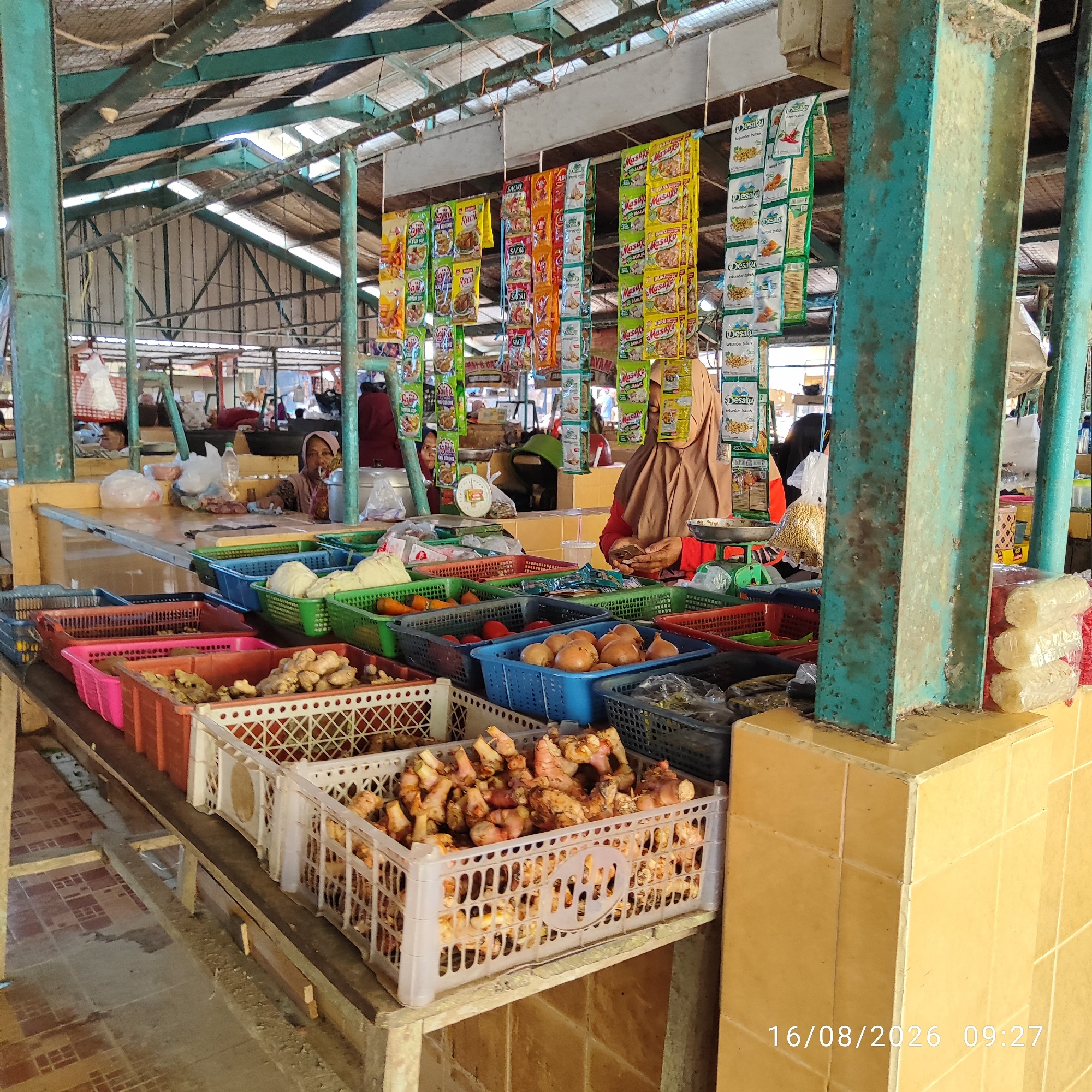 The image depicts an outdoor market stall in Pembaharuan, Indonesia. The stall is filled with various vegetables and spices displayed in plastic crates. In the foreground, white plastic crates overflow with ginger and other root vegetables. Behind these, various colored baskets hold onions, tomatoes, and what appear to be cabbages. Suspended from the market's roof are rows of packaged food items, likely seasonings or snacks, featuring colorful branding. Two women are visible behind the stall, one wearing a brown headscarf and red shirt, appears to be interacting with a mobile phone. The other woman, wearing a maroon headscarf, is partially obscured. The market structure is characterized by exposed metal beams and a corrugated metal roof, suggesting an open-air environment. The floor is tiled. A date and time stamp, "16/08/2026 09:27," is visible in the lower right corner.