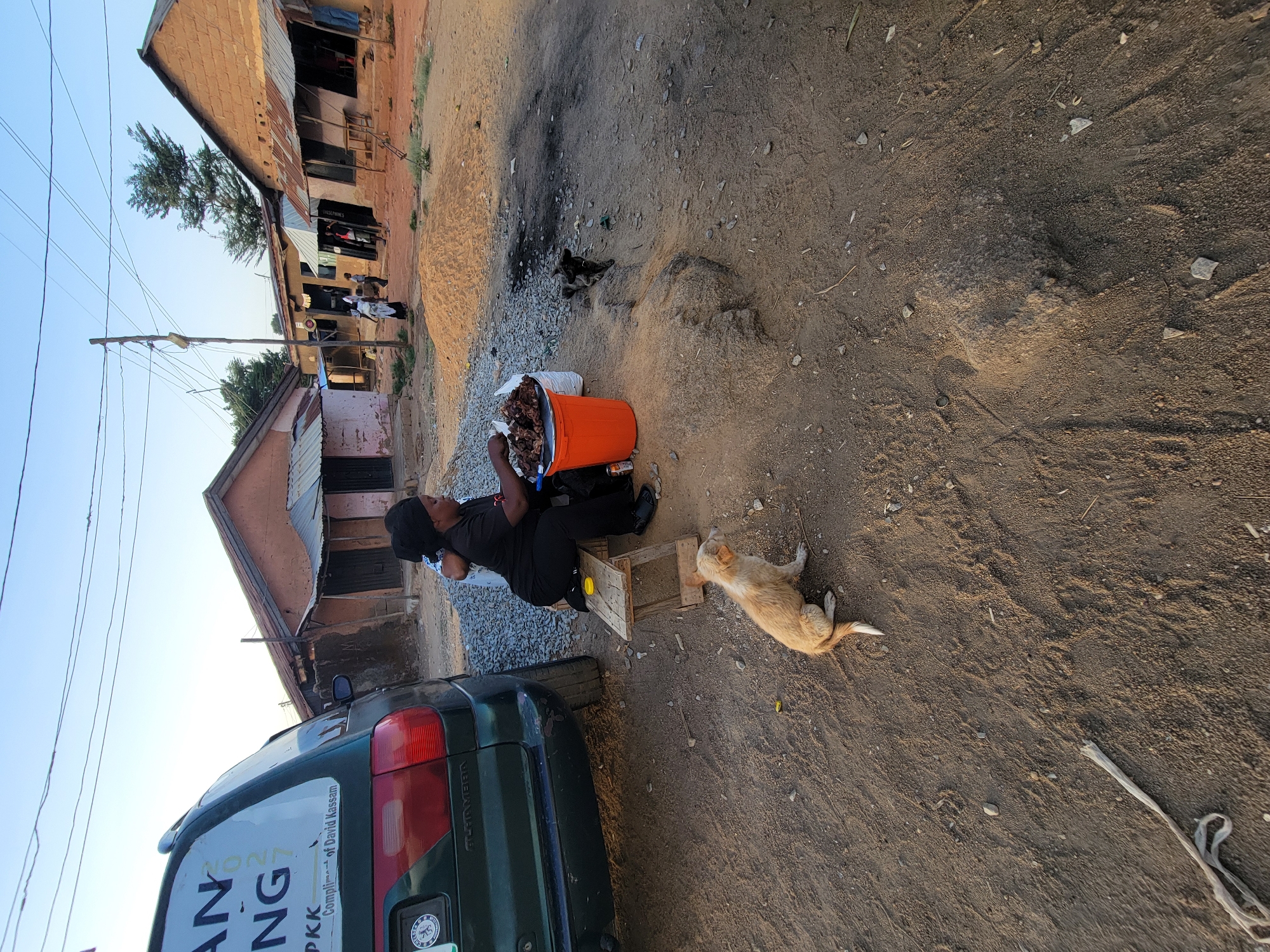 A woman sits on a wooden stool beside a green car on a dirt road in what appears to be Jos, Nigeria, based on the sticker on the vehicle. She is wearing black clothing and appears to be sorting through items in an orange bucket. A light-colored dog lies on the ground near the woman. In the background are buildings with corrugated metal roofs. Other people are visible further down the road. A pile of gravel and what appears to be burnt material are near the woman and the bucket.