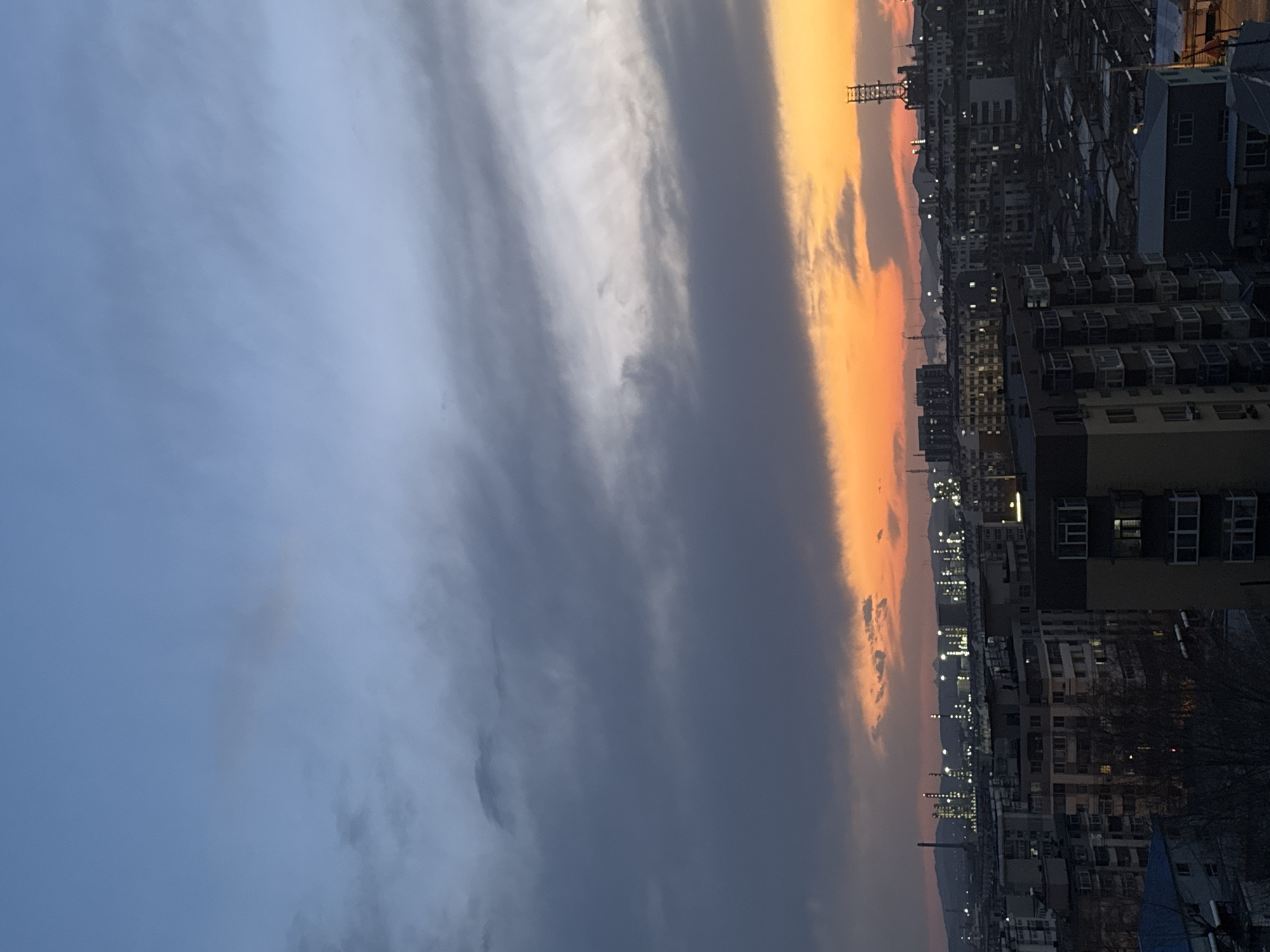The image shows an urban cityscape during twilight. The location is likely Zhongtun, China, though this is not definitively verifiable from the image itself.
On the right side of the image, multiple tall residential buildings are visible, densely packed together. Many windows are illuminated, suggesting it is late in the day. Utility poles are visible, and a transmission tower.
The sky occupies most of the image, with a gradient from dark grey/blue clouds on the left, to a vibrant orange and yellow sunset on the right. The horizon line is obscured by the buildings. No people are visible in the image.
