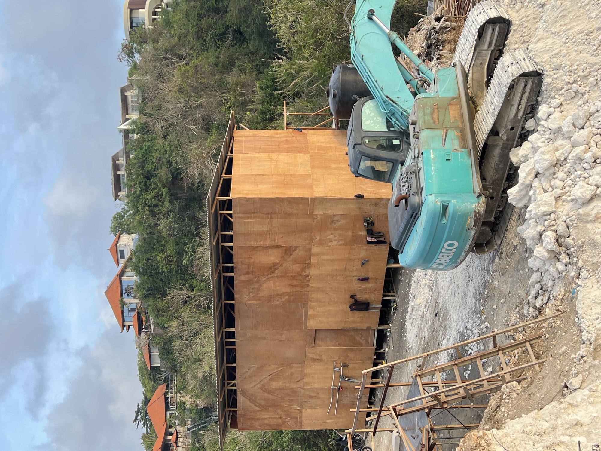 The image depicts a construction site located in Kuta Selatan, Indonesia, with a backdrop of hillside villas. In the foreground, a large, temporary wooden structure stands, appearing to be a shelter or storage facility, with laundry hanging on its exterior. A turquoise excavator, identified by the "KOBELCO" logo, is positioned in front of the structure, and next to it, a black water tank is visible. A wooden ladder leans against a scaffolding structure to the left of the wooden building. The ground is covered with debris, rocks, and construction materials. In the background, lush green vegetation covers the hillside, topped with several multi-story villas featuring red-tiled roofs. The sky above is partly cloudy.
