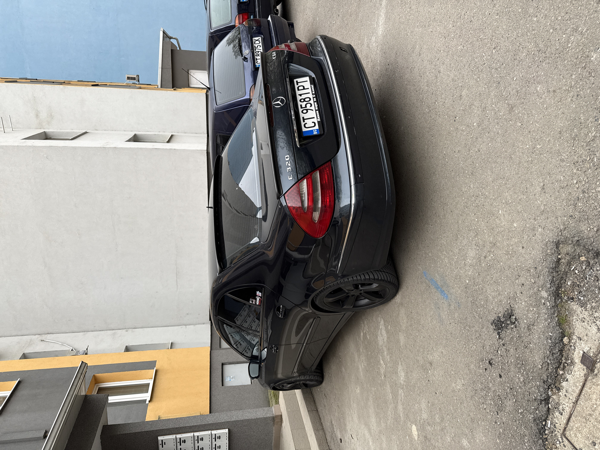 The image shows two parked cars on a street. The foreground car is a black Mercedes-Benz E320 with Bulgarian license plate CT 9581 PT, with a bumper sticker that translates to "I don't have money for 6 classes!" The car behind it is dark blue and has the license plate CT 8975 CK. The cars are parked next to a building with gray and yellow walls. There are mailboxes on the wall next to the Mercedes-Benz. The street is asphalt and has some stains and debris. The photo appears to have been taken in Stara Zagora, Bulgaria, given the Bulgarian license plates.