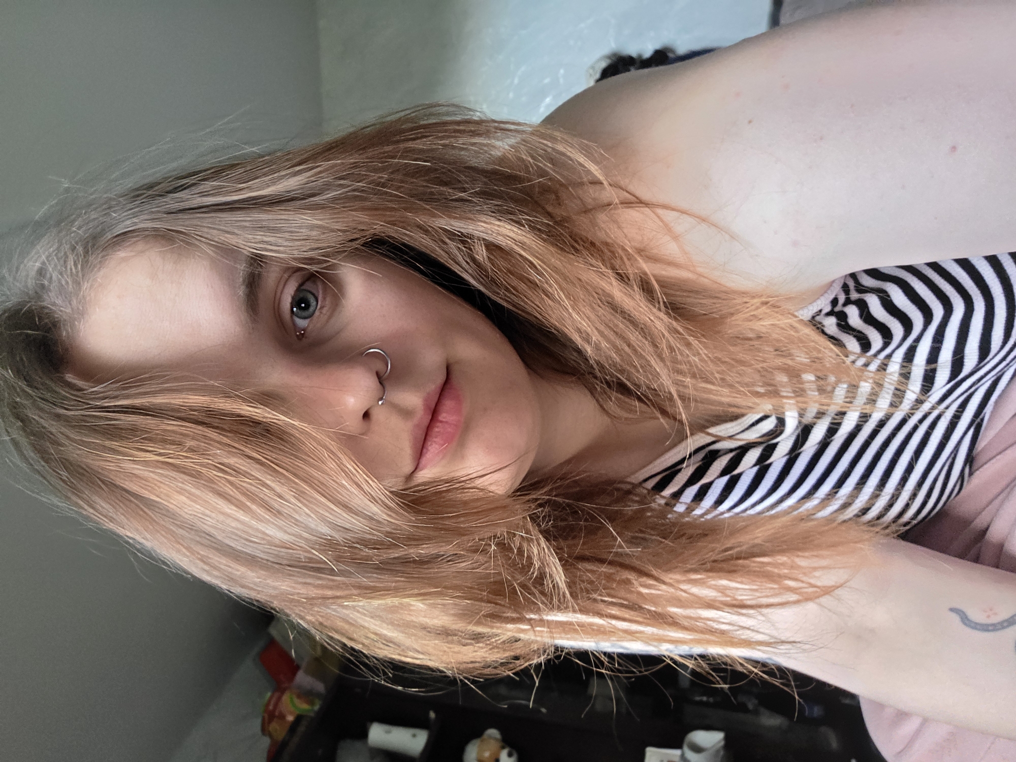 A close-up, angled, indoor photograph depicts a woman with light skin and shoulder-length, strawberry blonde hair. Her hair, partially obscuring her face, is lighter towards the roots. She has light blue eyes, a nose ring in her left nostril, and pink lips. She wears a black and white striped top and a pale pink garment is also visible. A glimpse of a tattoo is seen on her left forearm. The background includes a neutral-toned wall and a cluttered surface with various objects.