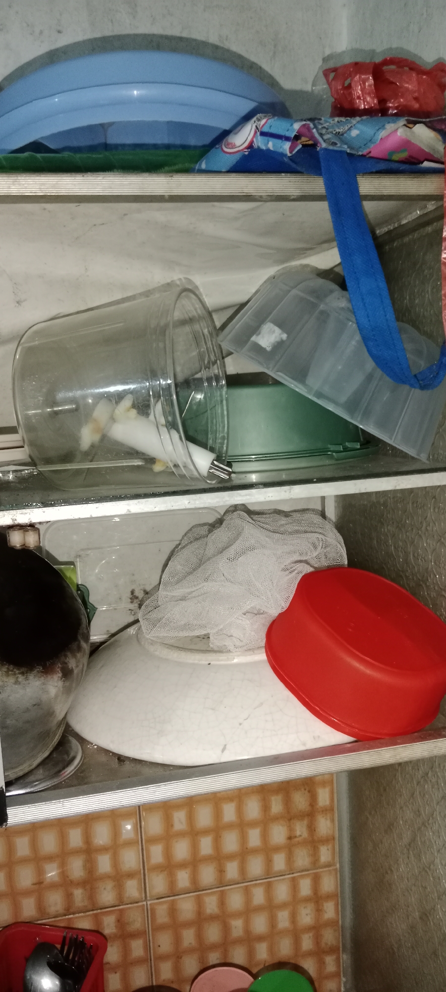 The image shows a cluttered shelf in what appears to be a kitchen or storage area. On the top shelf, a blue plastic basin is visible, along with a colorful cloth bag and a crumpled red plastic bag. The middle shelf holds a large clear plastic container that has tipped over, with a white object inside. Next to it is a green bowl or container, partially obscured by a clear plastic lid. On the bottom shelf, a large white plate with a crackled pattern is prominent. It is covered in part by a white mesh bag. To the right of the plate, a red plastic container lid is placed. To the left of the plate, a dark pot or container is visible. The background wall is textured and appears to be tiled. The overall scene suggests disarray and a lack of organization in the storage area. The location is identified as Subang, Indonesia, implying this is a scene from a residence or commercial establishment within that city.