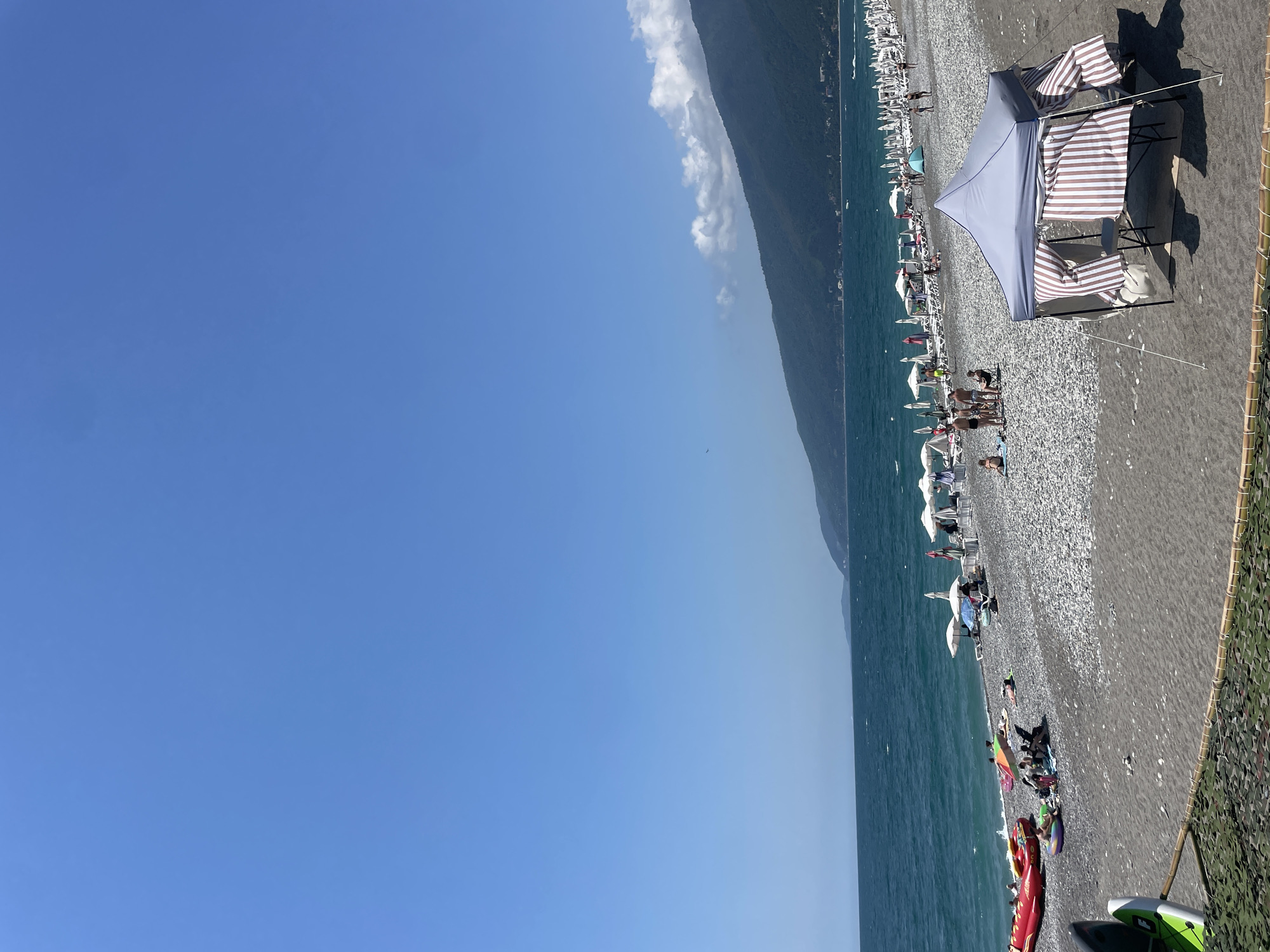 The image depicts a sunny day at a pebble beach in Gagra, Abkhazia. The sea is a vibrant blue-green, with gentle waves lapping against the shore. A line of white beach umbrellas and loungers stretches along the beach, some occupied by sunbathers. People are seen scattered across the beach, some sitting, some lying down, and some interacting in small groups. In the foreground, a grey pop-up tent with striped sides is set up, with a chair visible inside. Several inflatable water toys, including red and yellow tubes and a purple ring, are present near the water's edge. In the background, a lush green mountain range rises from the coast, with a few buildings visible on its slopes. The sky is a clear, bright blue with a few wispy clouds near the mountains. The overall scene suggests a relaxed and leisurely atmosphere at a popular tourist destination.