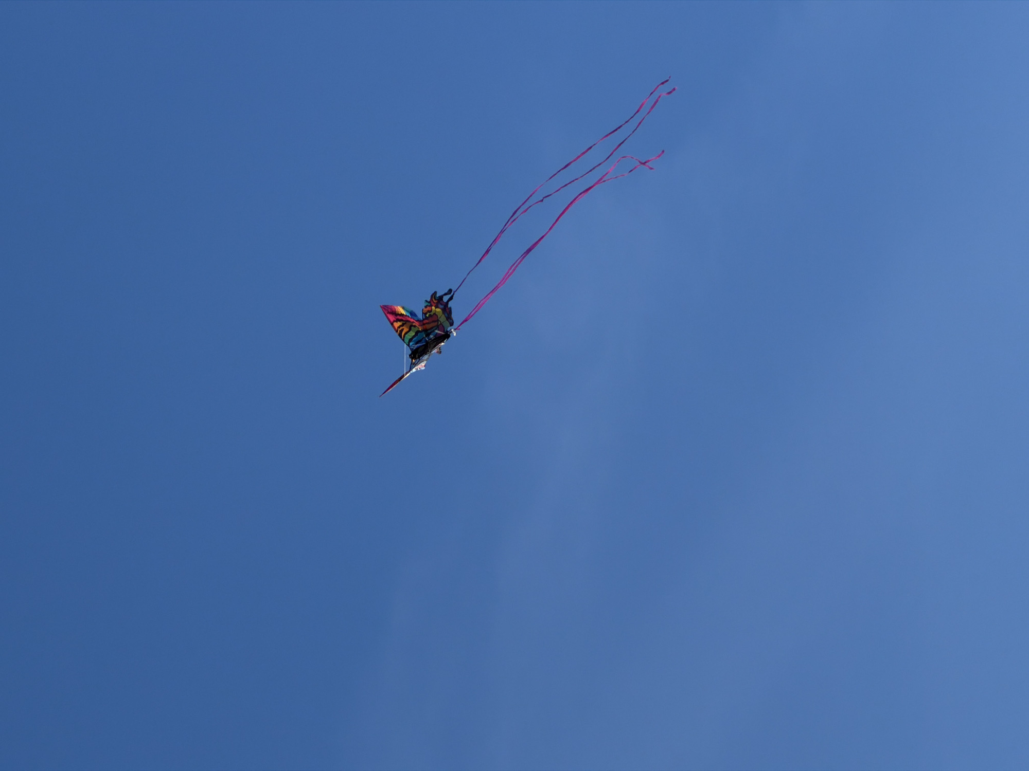 A colorful kite with streamers is flying high in a clear blue sky. The kite appears to be shaped like a butterfly or some other winged creature, and it has a long tail made up of three red streamers. The location of the city of Vancouver, Canada, cannot be determined from this image.