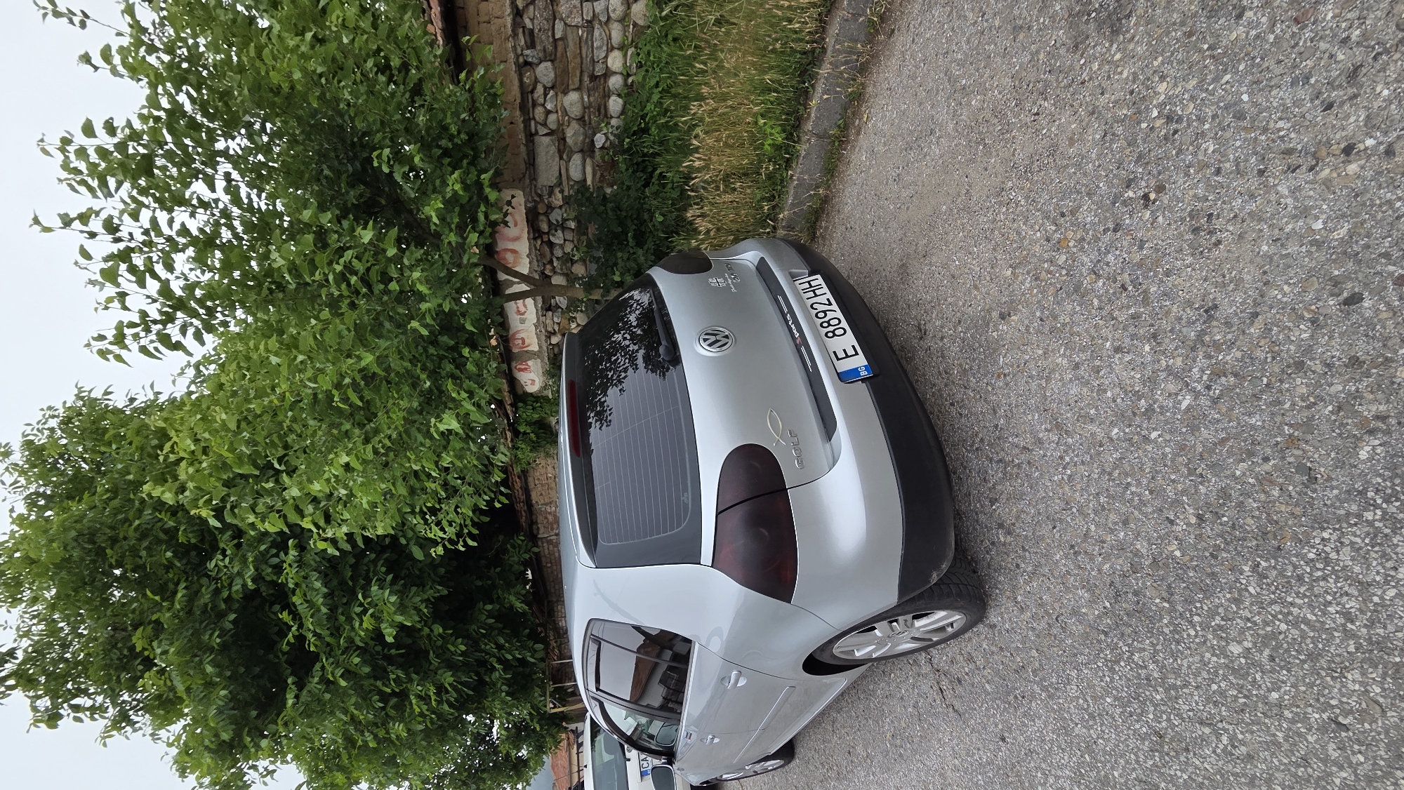 A silver Volkswagen Golf is parked on a gravel road next to a stone wall. The car is a hatchback and has tinted windows. The license plate reads "E 8892 HH". There are trees and grass growing in the background. The sky is overcast. The image appears to have been taken outdoors in a rural or suburban area.