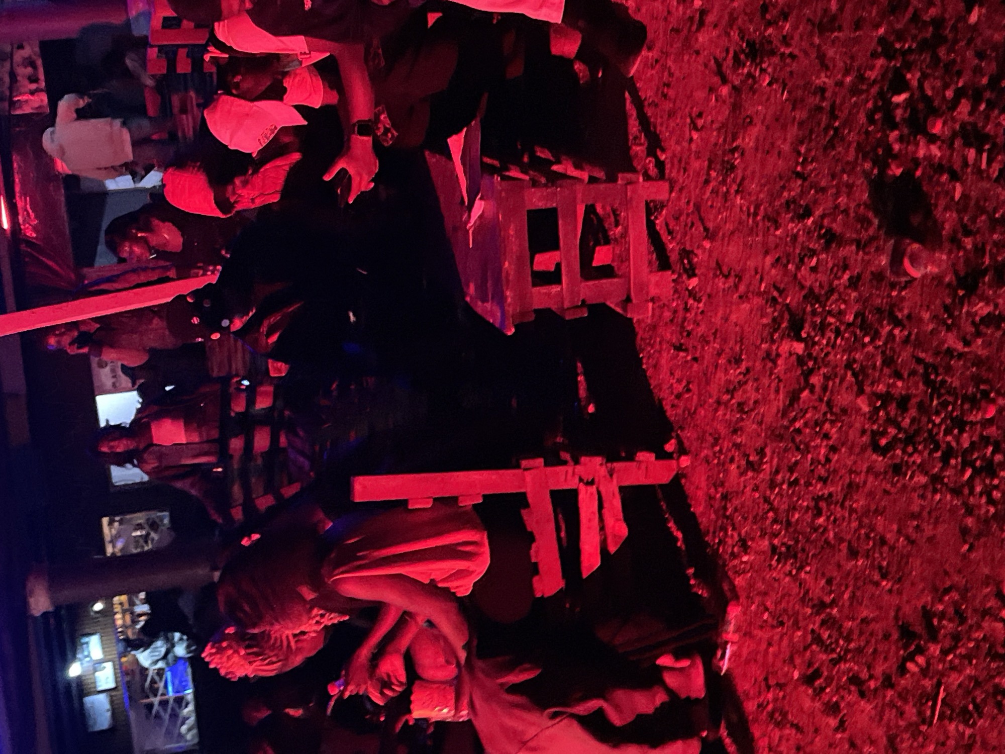 The image depicts a gathering of people in what appears to be an outdoor or semi-outdoor venue at night, illuminated by intense red lighting. The setting resembles a social event or party, possibly in a bar or club. In the foreground, a woman with braided hair is seated at a small table, holding a phone. To her right, a wooden chair is positioned. Further back, several other individuals are visible, some standing and others seated. One person is seen wearing a hooded garment, and another is wearing a light-colored hoodie. The ground is covered with gravel or small stones. The overall atmosphere suggests a lively and dimly lit social gathering. The specific location in Durumi, Nigeria, is not identifiable from the visual information provided.
