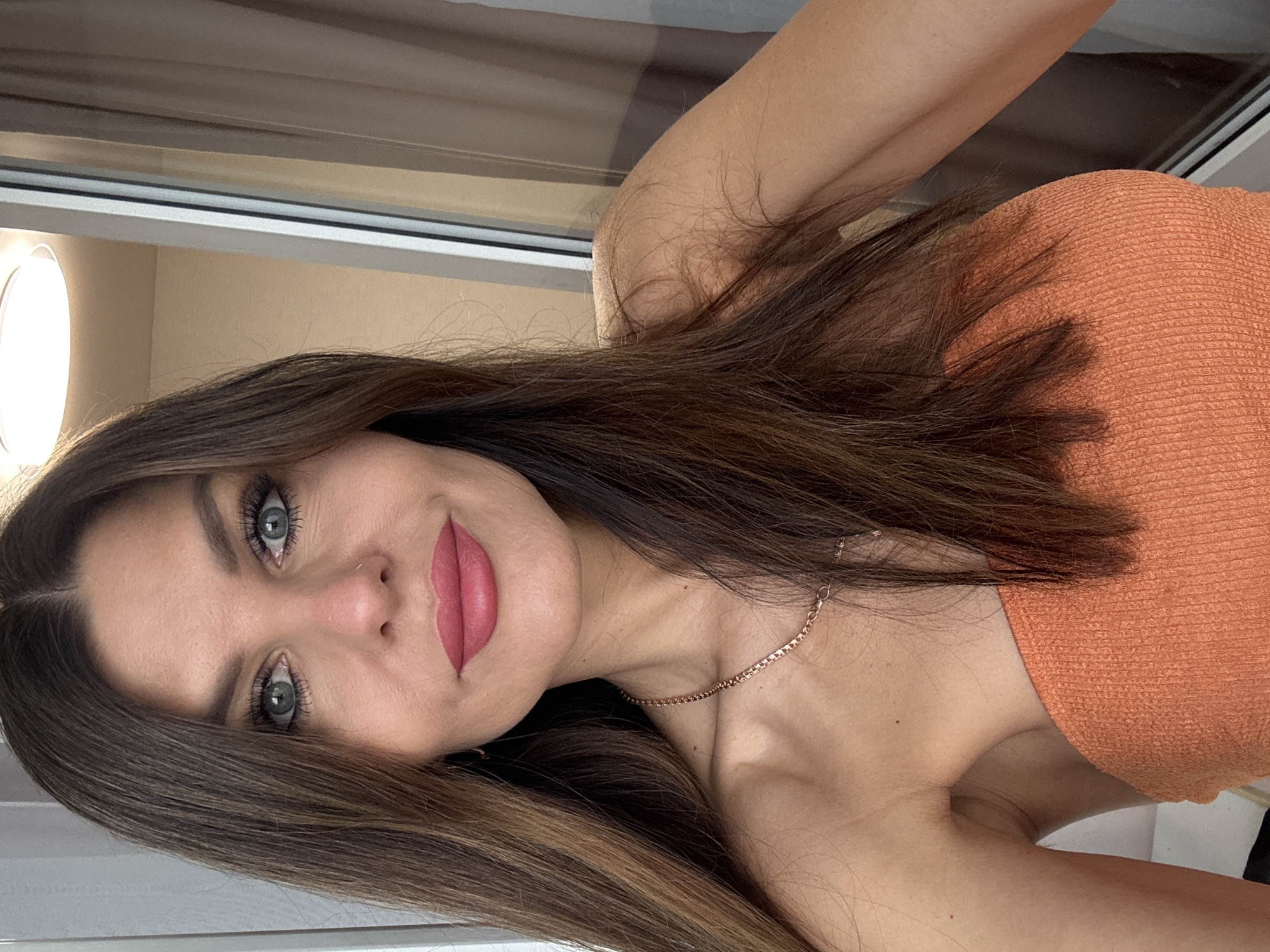 A medium-shot portrait captures a young woman indoors in Chaikovsky, Russia. The image is oriented with the woman's body appearing to be lying on her side, but her face is tilted to look directly at the camera. She has long, dark brown hair with lighter highlights, which is parted down the middle and flows around her shoulders.
Her face is the central focus, displaying large, light blue-green eyes enhanced with mascara and well-groomed eyebrows. She wears a small nose ring on her left nostril and has full lips colored with soft pink lipstick, forming a gentle smile. She appears to be wearing an orange, textured, off-the-shoulder top or dress. A thin, gold-colored chain necklace adorns her neck. Her bare arm is visible, bent upwards, suggesting a relaxed pose.
The background shows an indoor setting, likely a room with a window or a large glass partition along the top. A bright, circular light source is visible on the far left, possibly a lamp or natural light from an opening. The wall behind her is a light, neutral color. The overall mood is calm and engaging, as she looks directly at the viewer. There is no visible text in the image.
