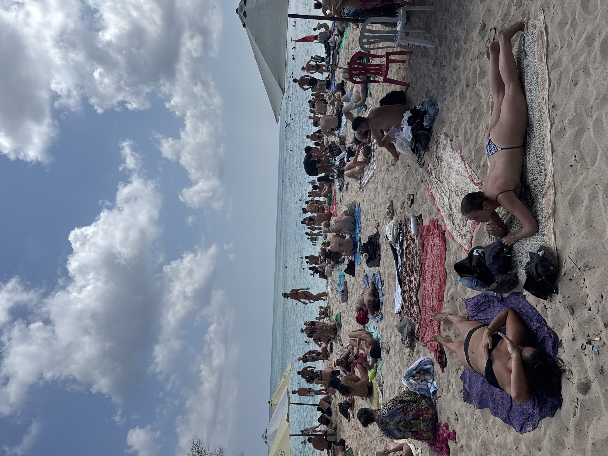 A crowded beach scene in Kuta Selatan, Indonesia, features numerous people sunbathing on the sand, some in swimwear, others in casual clothing. The beach is lined with umbrellas providing shade, and beach towels are scattered across the sand, indicating various resting and tanning spots. The ocean is visible in the background with several individuals swimming or wading in the water. The sky above is partly cloudy with a mix of blue sky and white clouds. Several beach chairs and bags are present, suggesting a day of leisure and recreation for the beachgoers.