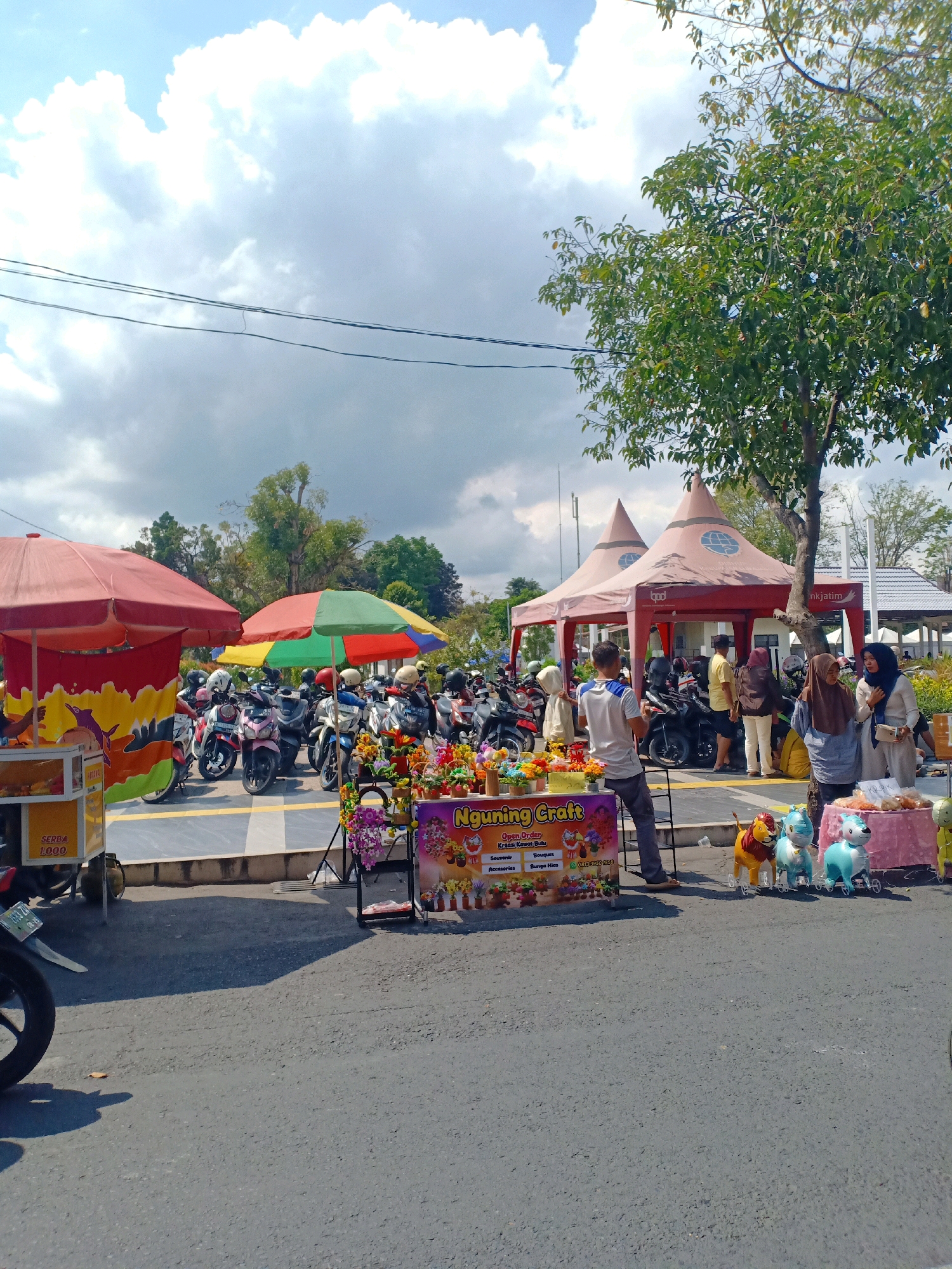 The image depicts an outdoor scene in Lumajang, Indonesia, with a focus on a marketplace area featuring several stalls and parked motorcycles. In the foreground, a paved road is visible, casting shadows from overhead objects. To the left, a food stall with a red umbrella displays items like "SERBA 1000" on its signage, suggesting it sells goods for one thousand units of currency. Adjacent to this, a colorful display of artificial flowers and other small crafts is set up.
Further into the scene, a row of parked motorcycles, some with helmets on, indicates a bustling area. Two large, conical tents, one red and one a lighter shade, are set up, possibly serving as shelter for vendors or as exhibition spaces. People are seen interacting in the area. A man in a white shirt and dark pants stands near the flower stall. Several women, some wearing hijabs, are clustered around another stall with a pink tablecloth draped over it, displaying inflatable animal toys, including a lion and two horse-like figures. One woman is holding a white bag, and another appears to be engaged in a transaction. The background reveals more trees, buildings with tiled roofs, and a cloudy sky with sunlight breaking through. The overall atmosphere suggests a typical daytime scene in a local market or gathering place in Lumajang.