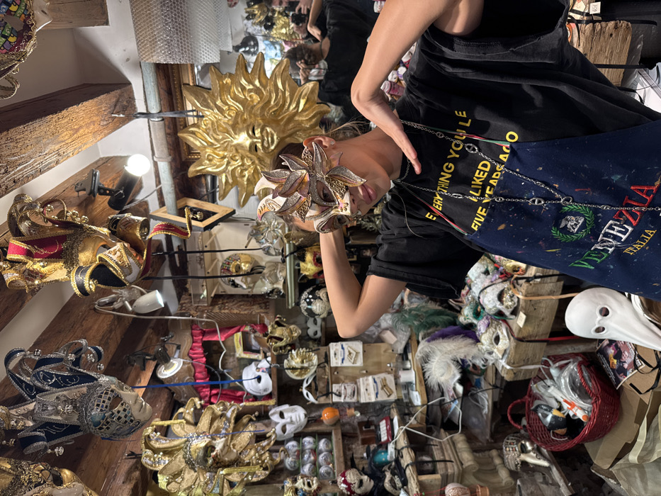 An indoor scene depicts a retail establishment, specifically a Venetian mask shop in Venice, Italy. In the foreground, a person with light-colored hair, wearing a black t-shirt with yellow text partially visible as "EVERYTHING YOU LE," and a paint-splattered blue apron embroidered with "VENEZIA ITALIA," poses while wearing an ornate, light-colored half-face Venetian mask adorned with gold and bronze leaf-like motifs. Their arms are raised, with hands positioned behind their head, slightly adjusting the mask. A long silver chain necklace is draped over their shirt and apron.
The shop interior is densely packed with merchandise. Numerous Venetian masks of various styles, colors, and materials hang from rough wooden ceiling beams and are displayed on walls, shelves, and tabletops. Prominent hanging masks include multi-pointed jester-style masks with bells (e.g., silver and black, red and gold variations). A large golden sun-face mask with radiating rays is mounted on the wall behind the main subject, to the right. On a table in the lower-left foreground, a white, long-nosed Plague Doctor mask is visible. Various other masks, some with feathers and intricate designs, are arranged on shelves and surfaces throughout the cluttered space.
The environment features exposed wooden ceiling beams, packed shelves, and tables laden with masks and other small items. Small light fixtures are visible. On the right side of the image, a reflection in a mirror shows another individual, holding a phone, suggesting a photographic interaction.