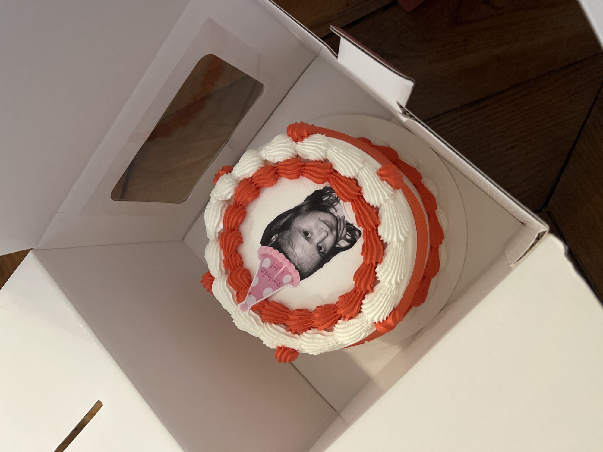 The image shows a round birthday cake inside an open white cardboard box. The cake has white frosting with red decorative piping along the edges. A black-and-white photograph of a child's face is placed in the center of the cake, topped with a small pink party hat graphic. The background reveals part of a wooden floor. The location is not indicated in the image. The city of Paris, France cannot be assumed.