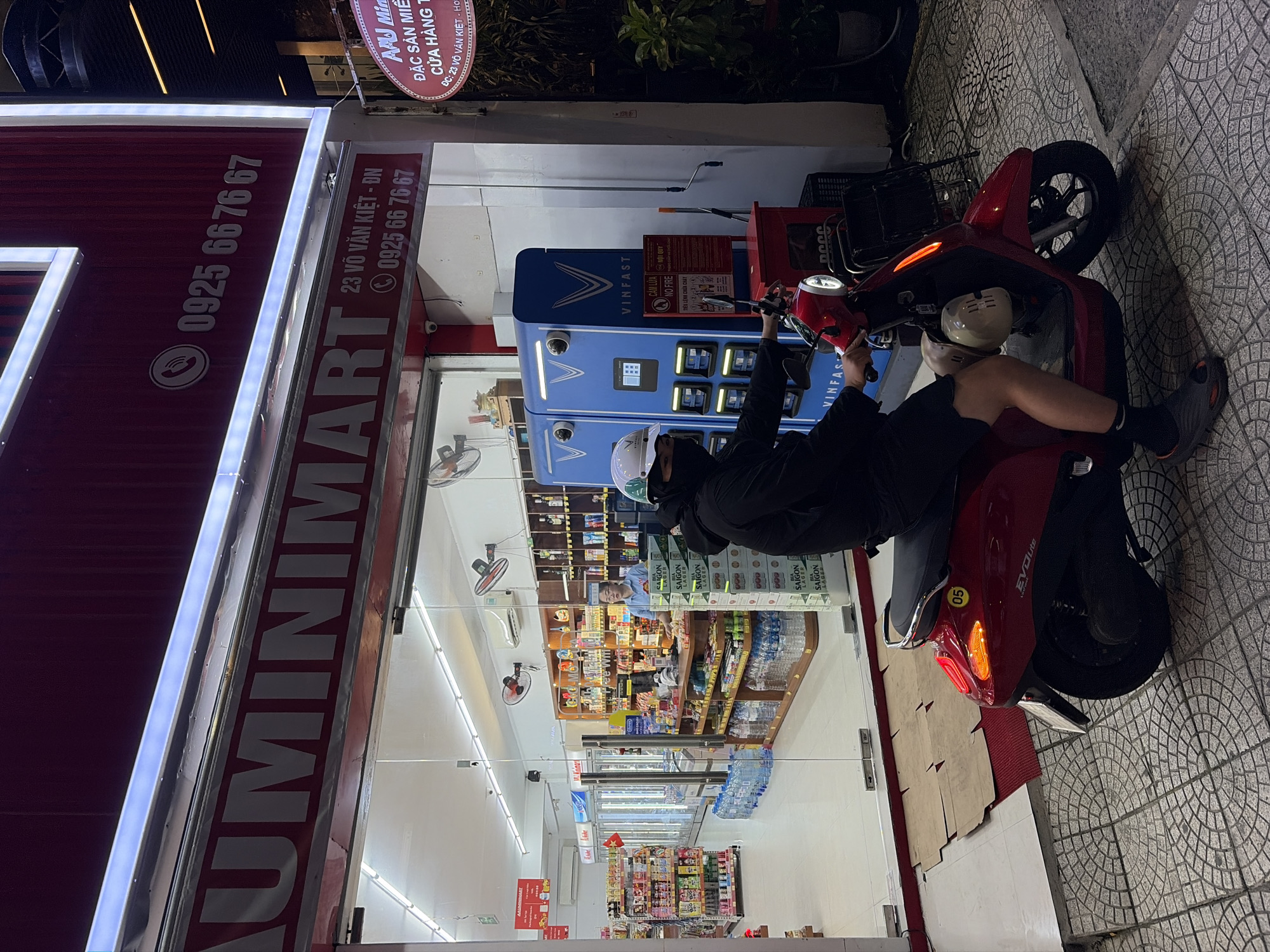 A person wearing a black hoodie, black shorts, and a white helmet is sitting on a red electric scooter parked on a tiled sidewalk in front of a convenience store called "AUMIMIMART" at night. The store is brightly lit, with shelves of products visible inside, including beverages and snacks. A VinFast charging station is located next to the store's entrance. The store sign displays the address "23 Võ Văn Kiệt - Đà Nẵng" and a phone number "0925 66 76 67". The scooter has a yellow sticker with the number "05" on its rear.