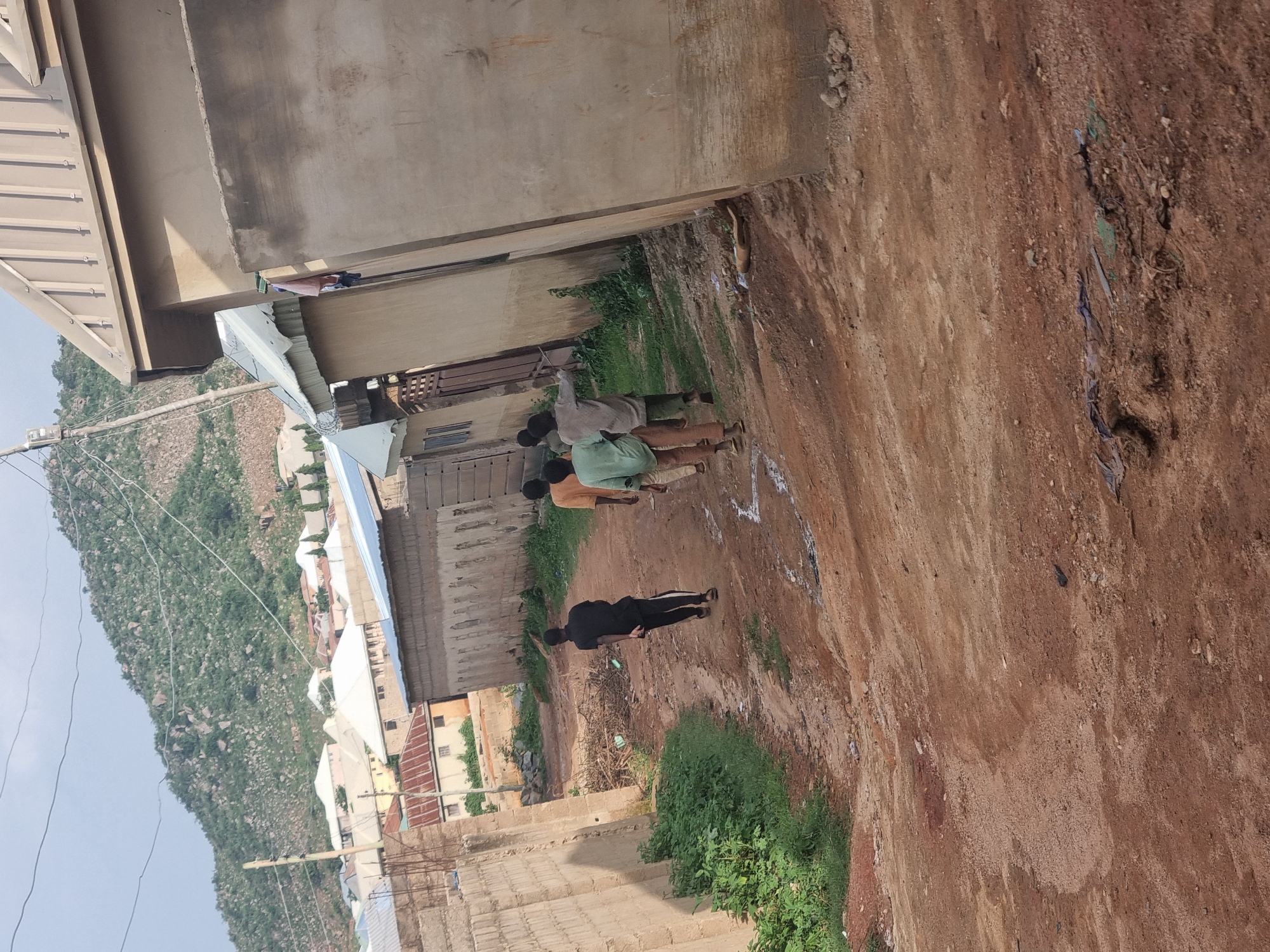 In Jos, Nigeria, a dirt path winds through a residential area, leading towards a steep, rocky hill covered in sparse vegetation. The path is flanked by simple, unplastered brick walls on one side and by a light-colored building with a corrugated metal roof on the other. Further down the path, several young boys are walking away from the camera. One boy in a black t-shirt and dark pants is in the foreground, while three others, dressed in light-colored shirts and pants, follow behind. The buildings in the background are of various designs and colors, with some featuring corrugated metal roofs and others with flat, concrete ones. The overall impression is of a developing urban area with a strong connection to the natural landscape.