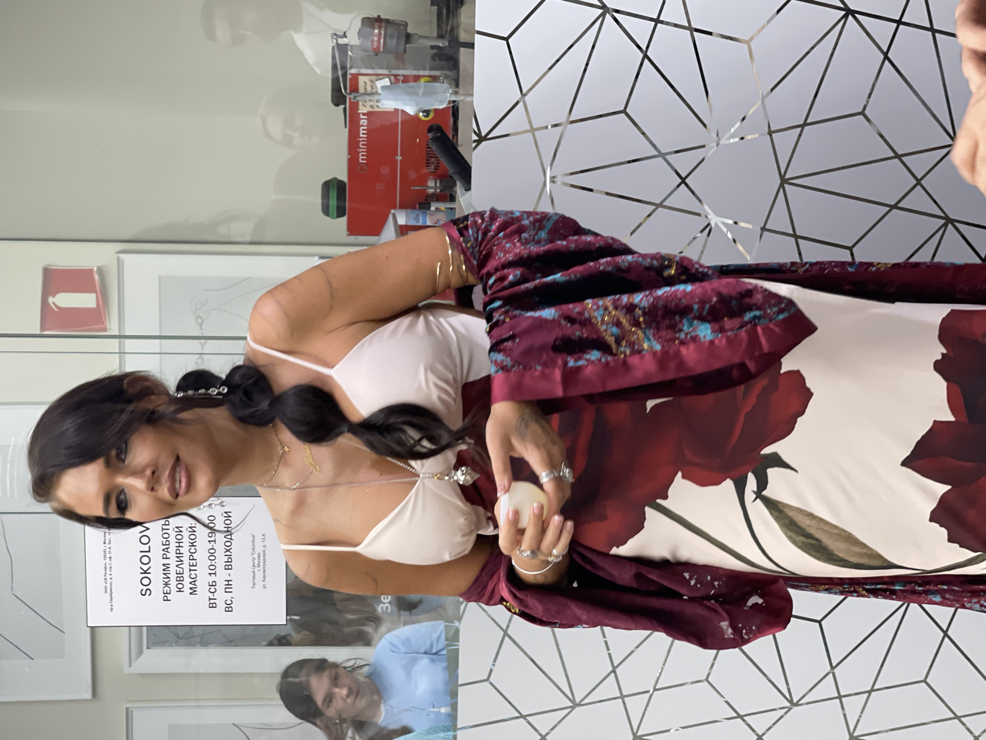 A woman stands in a detailed interior view of the Sokolov Jewelry Workshop located within the Columbus Shopping Center at Kirovogradskaya Street, 13-A, Moscow, Russia. The central figure, a woman in her 30s or 40s with dark hair braided over her left shoulder, looks slightly to her right with a gentle smile. She wears a white spaghetti-strap top, a long white dress adorned with large red floral patterns, and a burgundy shawl with intricate blue and gold detailing. Her visible accessories include a "Selena" nameplate necklace, a longer silver chain with an elaborate pendant, a dangling earring, multiple rings on both hands, and bracelets on her arms. She holds a small, translucent, light-colored cylindrical object in her cupped left hand. Behind her, a white sign prominently displays "SOKOLOV ЮВЕЛИРНОЙ МАСТЕРСКОЙ" (Sokolov Jewelry Workshop) with operating hours "ВТ-СБ 10:00-19:00" (Tuesday-Saturday 10:00-19:00) and "ВС, ПН - ВЫХОДНОЙ" (Sunday, Monday - CLOSED), explicitly stating the address as "Торговый Центр "Columbus", г. Москва, ул. Кировоградская, д. 13-А". The foreground is partially obscured by a glass panel featuring a geometric, reflective pattern. Through the glass, a young woman is visible to the left, and reflections on the right show a red "minimart" machine, industrial equipment, a fire extinguisher sign, and blurred figures of other individuals.