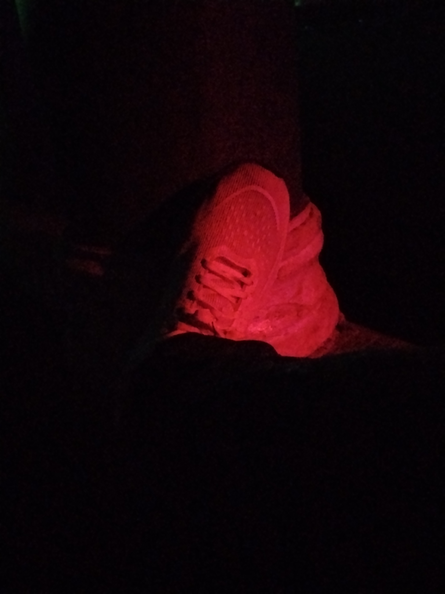 In Durumi, Nigeria, a close-up shot reveals a pair of red sneakers illuminated by a red light, creating a dramatic and shadowy effect. The shoes are positioned on what appears to be a dark surface, possibly a floor or ground. The lighting casts deep shadows, obscuring much of the surrounding environment, but the texture of the shoe material is visible, suggesting a mesh or fabric construction. The scene is dimly lit, with only the shoes being the primary focus due to the strong red illumination.