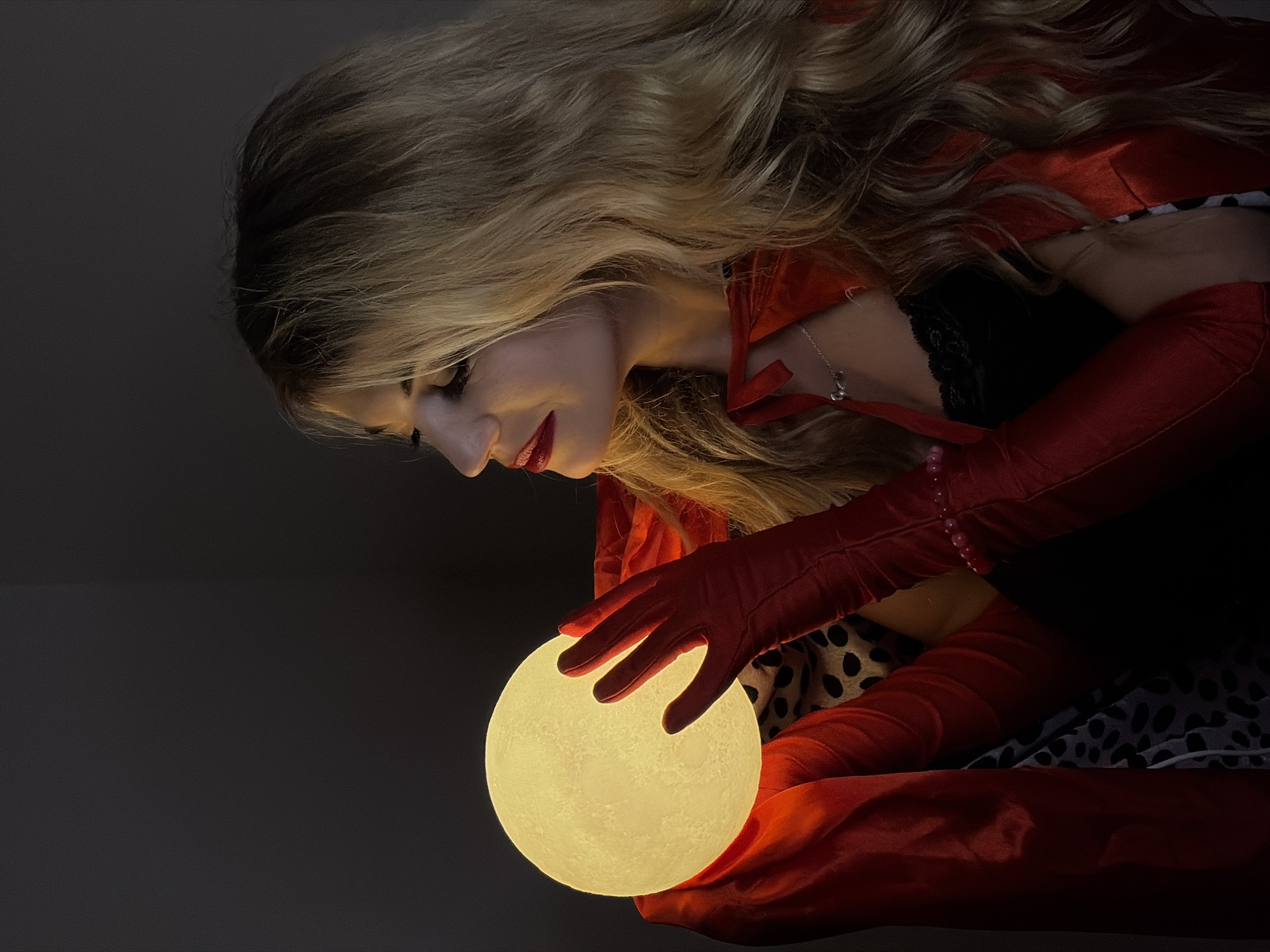 A woman with blonde hair and red lipstick is holding a glowing orb. She is wearing red gloves and a red garment, possibly a cape, over a black top with lace trim and a black and white patterned dress or skirt. She is looking down at the orb, which is the primary light source in the image. The background is dark, suggesting a dimly lit room. It is impossible to verify whether this image was taken in Budva, Montenegro or to know any specific location within a city.