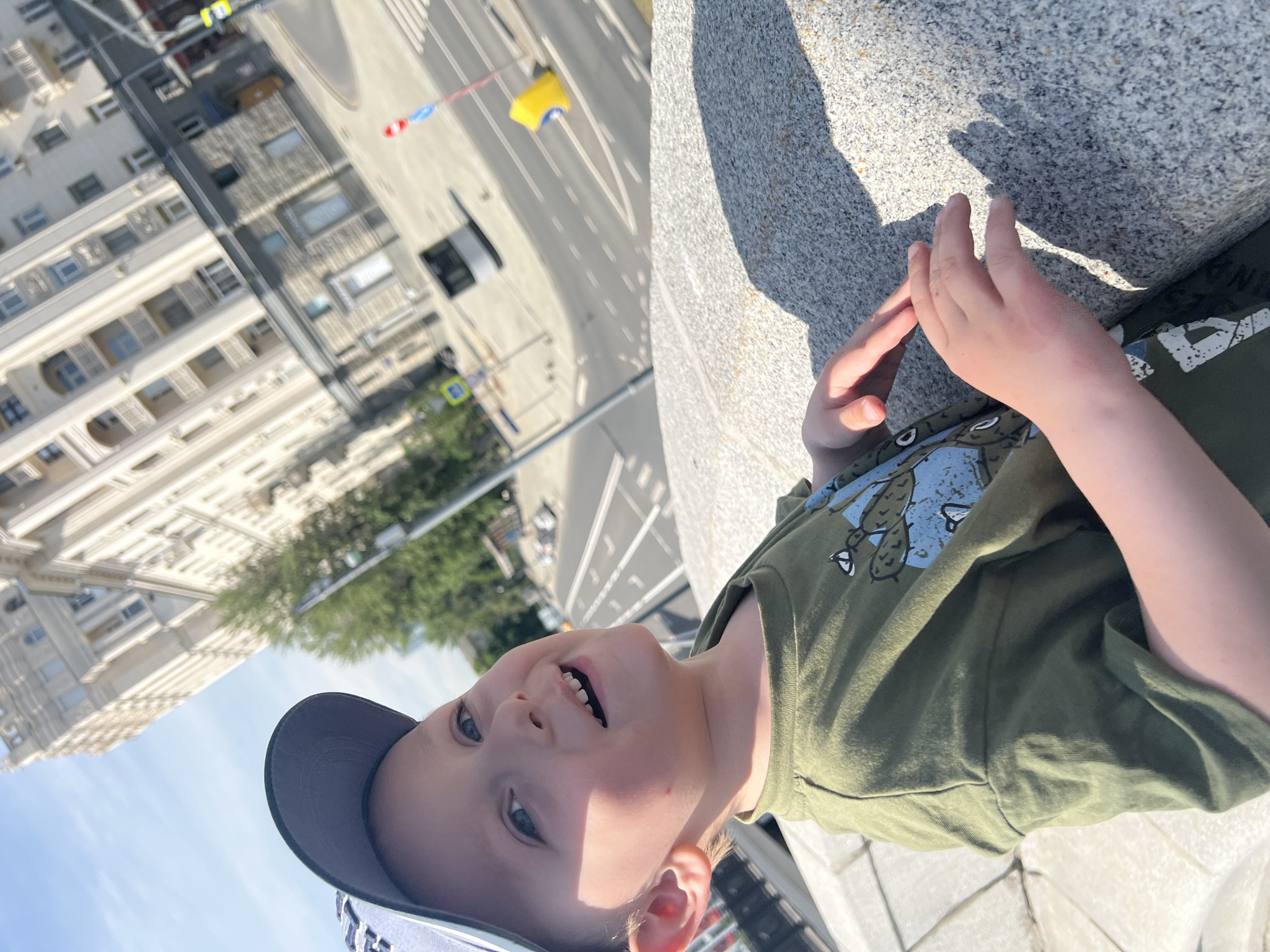 The image depicts a young boy in the foreground, positioned in an outdoor urban setting during daylight under clear skies. He is wearing a blue-grey baseball cap and a green t-shirt with a light blue graphic design featuring two cactuses and additional white details. The boy, with fair skin and light hair, is smiling broadly, revealing his teeth, and looking slightly upwards and to the viewer's left. His right hand rests on a grey, speckled stone parapet or wall that occupies the lower right portion of the frame. Strong sunlight casts distinct shadows on this surface.
In the midground, a multi-lane asphalt road with white painted markings (dashed lines, solid lines, crosswalk) is visible. A dark-colored bus is partially seen on the road. Various street elements are present, including a yellow, potentially plastic, traffic delineator or barrier on the right side of the road, a red and blue circular traffic sign on a pole, and a blue square directional sign further down on the left. Green trees are interspersed between the street and the background buildings.
The background is dominated by a grand, multi-story building of light-colored stone or concrete, featuring numerous windows, balconies, and classical architectural details, characteristic of Stalinist Empire style. A smaller, darker stone-faced building is situated below it at street level. This prominent building is the Radisson Collection Hotel, Moscow (formerly Hotel Ukraina), one of the "Seven Sisters" skyscrapers. The photograph is taken from an elevated position, looking down onto Kutuzovsky Prospekt and across towards the hotel, indicating the vantage point is likely from the Borodinsky Bridge over the Moskva River in Moscow, Russia.
