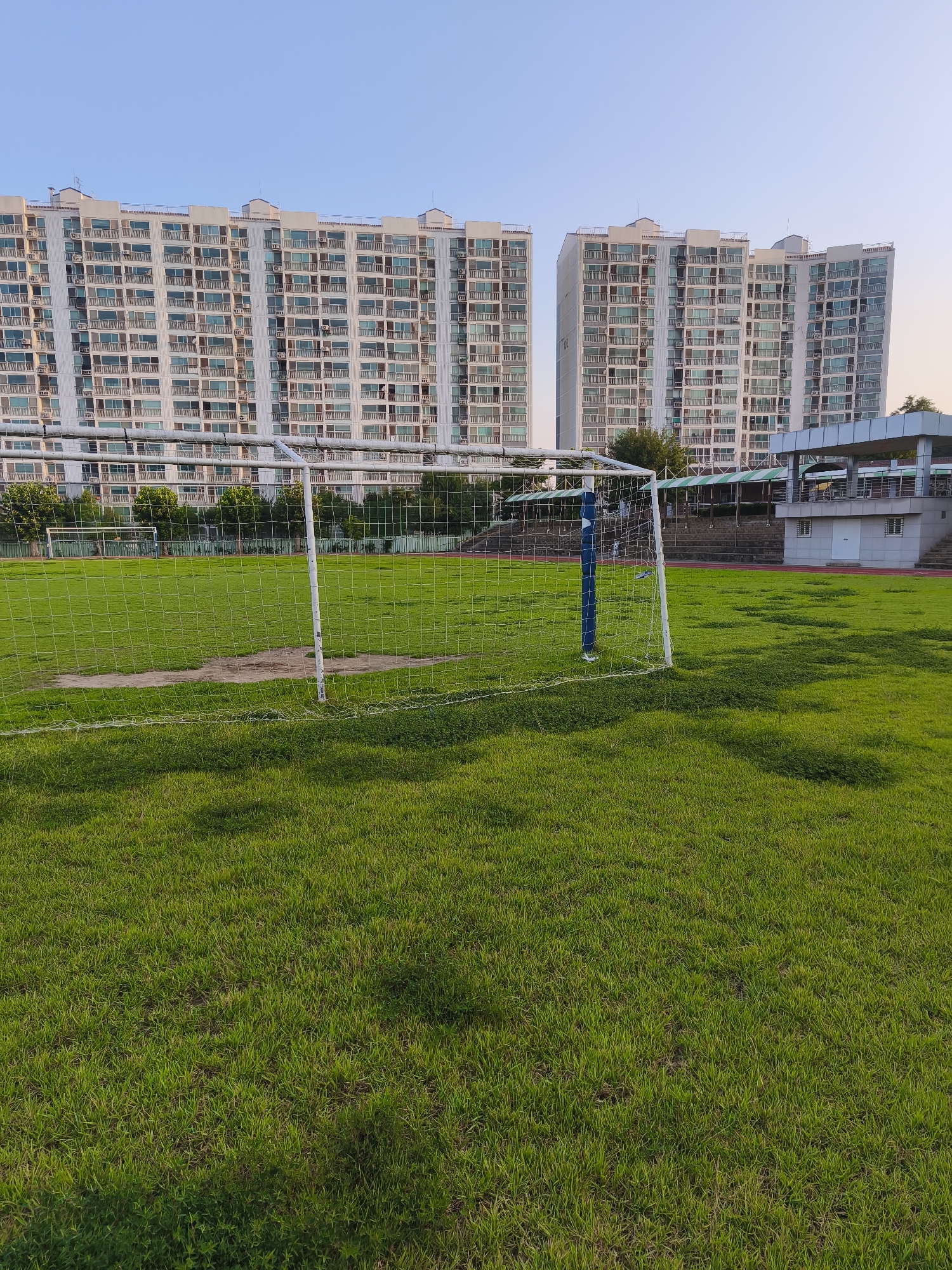 The image depicts a view of a grassy field with a soccer goal in the foreground. In the background, two large, multi-story apartment buildings with numerous windows and balconies are visible under a clear sky. To the right of the apartment buildings, a structure with a covered walkway and a small building is present. The scene appears to be in a residential area, likely a park or sports facility within the city of Asan-si, South Korea, given the architectural style of the buildings and the presence of a soccer field. The grass is green, indicating a well-maintained outdoor space. No people are visible in the image.
