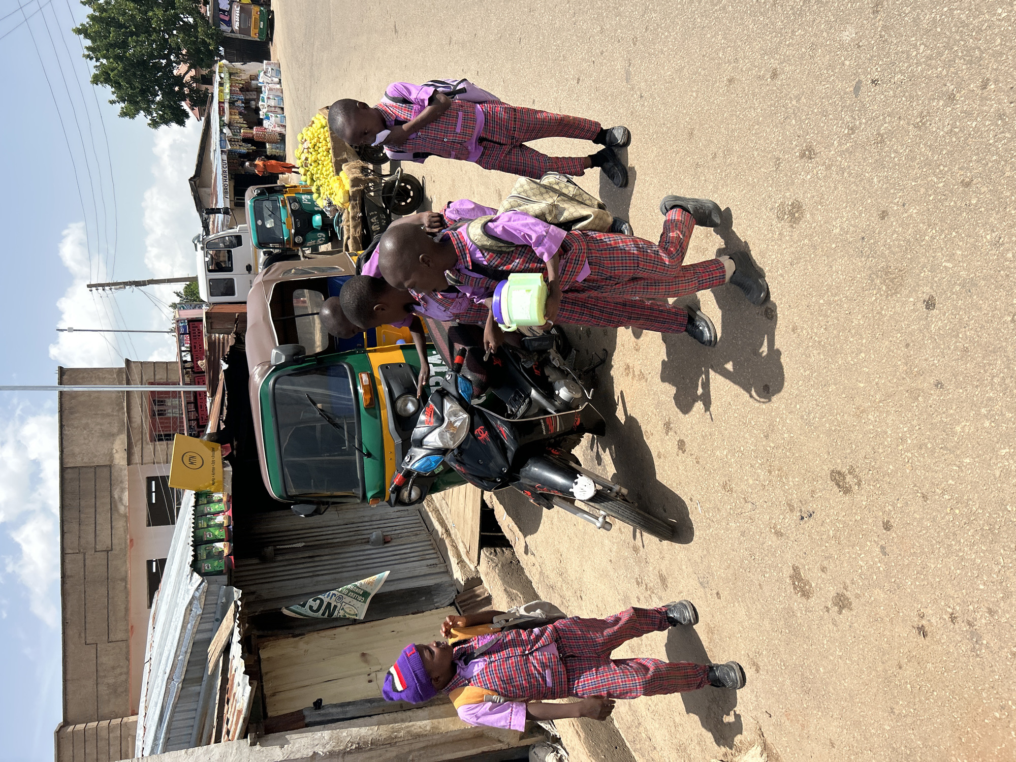 In Jos, Nigeria, five young boys in school uniforms stand around a motorcycle. The boys are all wearing plaid pants and light purple shirts, with some wearing vests. One boy wears a purple beanie. The street is paved with asphalt, and there are buildings and market stalls in the background. A tuk-tuk is parked behind the motorcycle, and a cart filled with yellow fruit is also visible. The sky is blue with scattered clouds. The scene appears to be a typical street in a bustling Nigerian city.