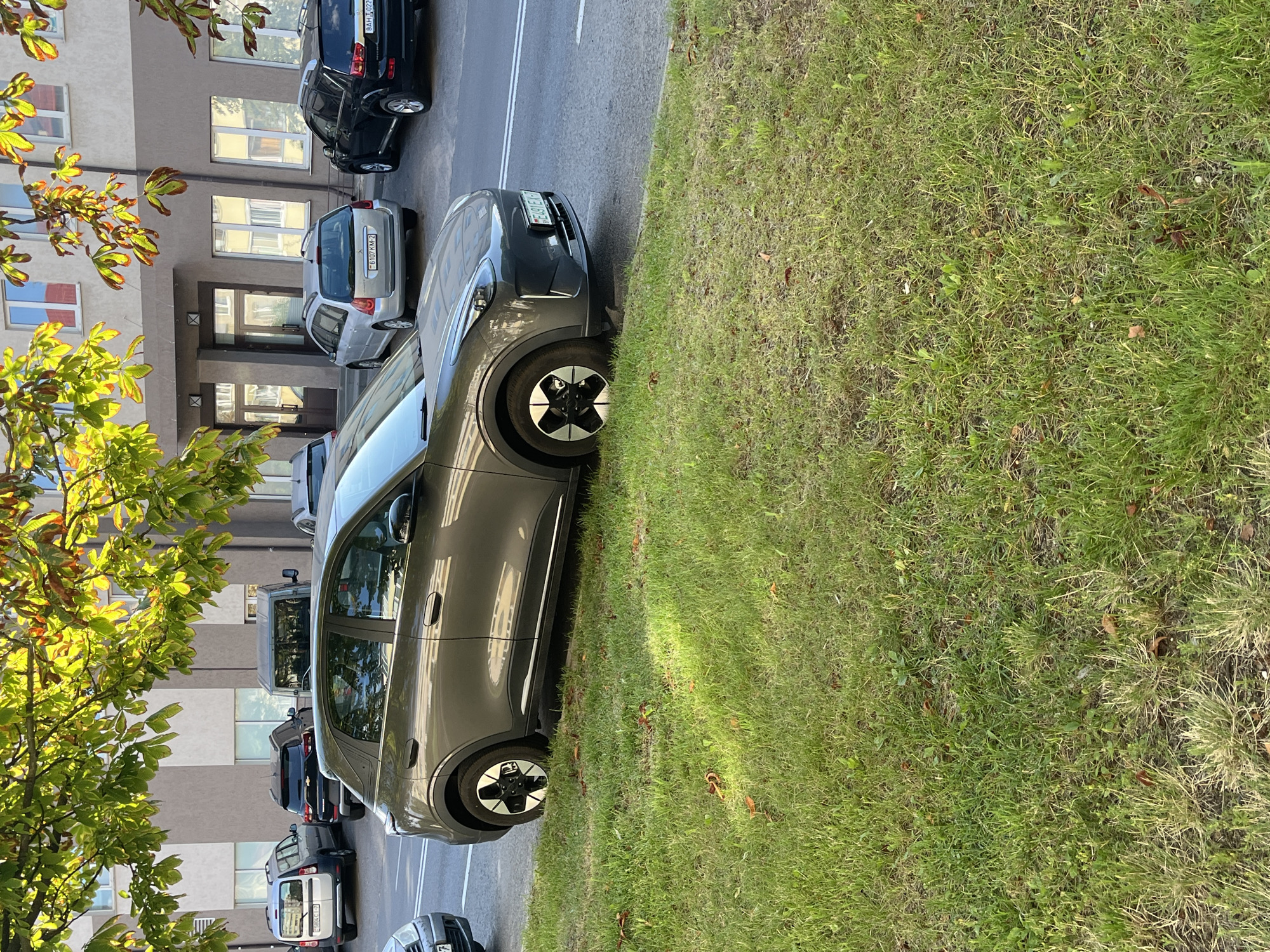 In Minsk, Belarus, an overcast, daytime scene depicts a grey hatchback parked on the grass verge adjacent to a paved road. The car, a compact SUV with distinctive futuristic styling, features unique black and silver wheels and a front license plate reading "AE491EX-7". Behind the hatchback, several other vehicles are visible parked on the road, including a black SUV and a silver station wagon. Further in the background, a light-colored building with multiple windows stands. The foreground is dominated by a lush green lawn, with a few fallen leaves scattered across it. The overall impression is one of an ordinary urban parking situation, possibly in a residential or commercial area of Minsk.