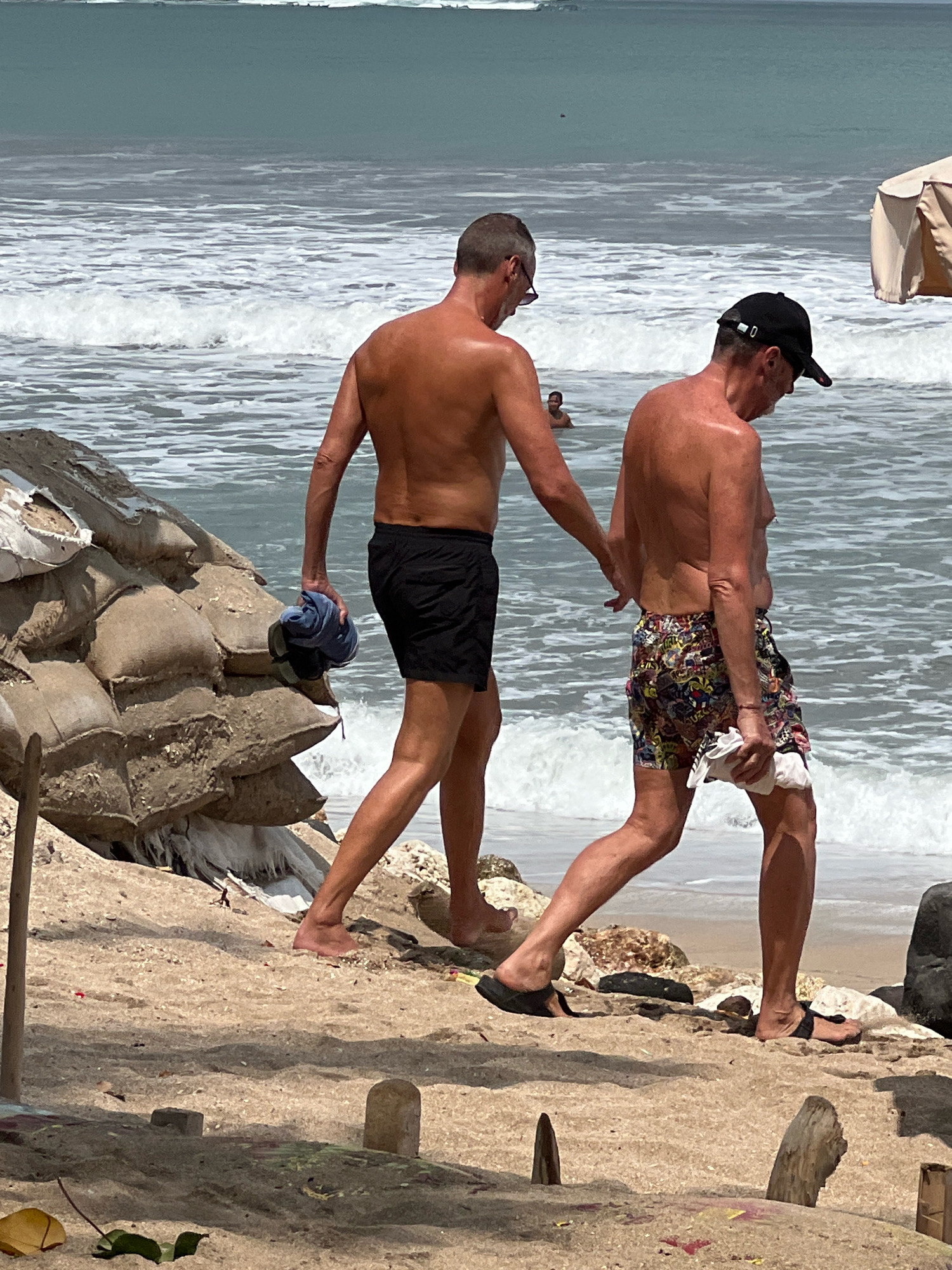 Two shirtless men walk along the sandy beach of Kuta, Indonesia, near the ocean. The man on the left, wearing black swim shorts and sunglasses, carries a blue towel and a pair of black flip-flops in his left hand. The man on the right, wearing a black baseball cap and colorful patterned swim shorts, carries a white towel in his right hand. In the background, the ocean has moderate waves, and a light-colored beach umbrella is visible. A pile of sandbags, likely part of a barrier, is to the left of the men on the beach. The foreground shows sand, small rocks, and scattered debris.