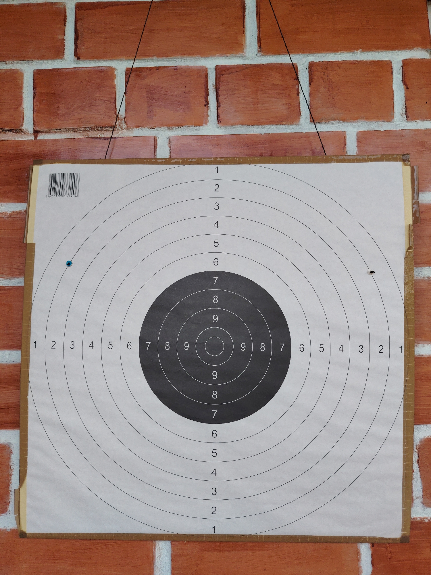 A shooting target is displayed against a brick wall. The target is a standard circular design with concentric rings numbered from 1 to 10. There are two visible bullet holes on the target, one slightly to the left of the center and another to the right. The target appears to be made of paper and is attached to the brick wall with tape. The brick wall is made of red bricks with white mortar. The image is taken from a medium distance, showing the entire target and a portion of the wall. The scene is static and does not depict any people or action. The location in Konakovo, Russia, is not visually evident from the image.
