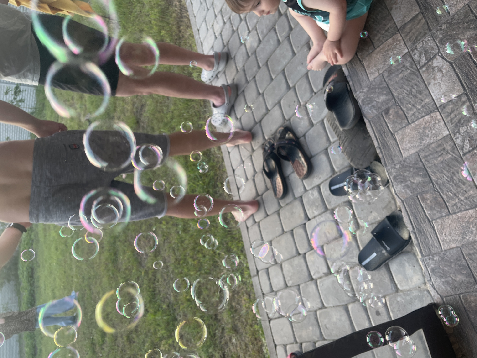 The image, captured in Petrivka, Ukraine, presents an outdoor scene characterized by numerous iridescent soap bubbles floating across the frame. The ground transitions from grey rectangular paving stones in the foreground and midground to green grass in the background. A white, horizontally paneled structure, possibly a fence or building, is visible behind the grass.
Four individuals are present. In the far left background, a person, facing away, wears a dark patterned top and light blue wide-leg trousers, standing on the grass. To their right, an adult male, bare-chested in grey athletic shorts with a small red logo, stands barefoot on the paving stones, facing slightly right. Adjacent to him, another adult male wears a light grey t-shirt and dark shorts, holding a yellow cloth in his right hand, standing on the paving in grey Crocs-style clogs, facing slightly left. In the right foreground, a young child, barefoot and clad in a teal and black outfit, sits on the paving stones, facing left and observing the bubbles.
Various items of footwear are scattered on the paving, including two black slide sandals and one pair of dark brown/black thong sandals. A black fabric object is partially visible in the bottom left corner. The scene is illuminated by diffuse daylight, emphasizing the vibrant reflections on the bubbles.
