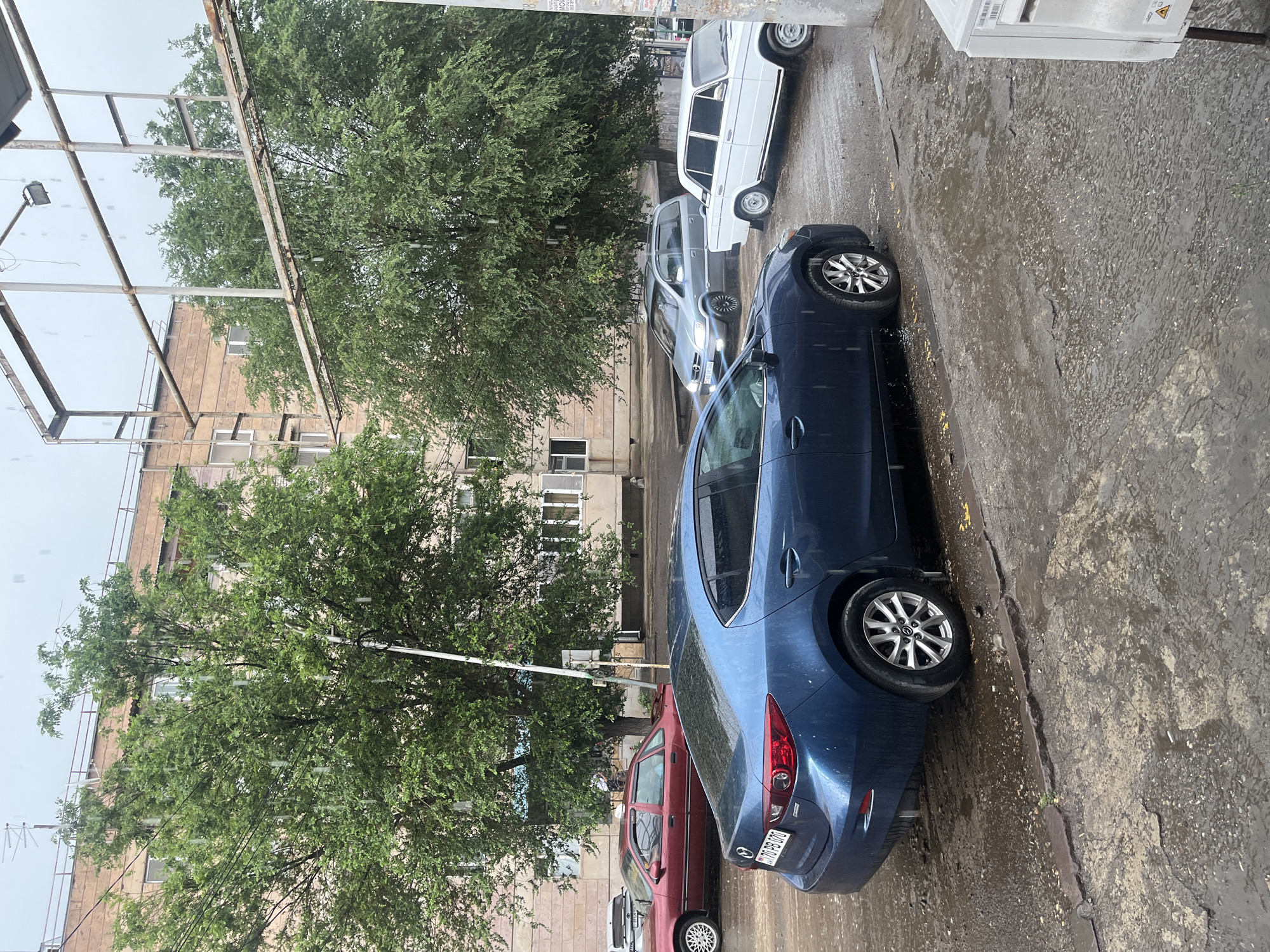 The image shows a street scene with several cars parked along a wet asphalt road. The weather appears to be rainy, with visible raindrops in the air and puddles on the ground. A dark blue sedan is prominently featured in the foreground, its taillight and rear wheel visible. Behind it, a red sedan is parked. Further down the street, a white sedan and a silver minivan are also visible.
The background includes a multi-story building with a light-colored facade and numerous windows. Lush green trees with full foliage line the street, suggesting a warm season. Overhead, a metal structure, possibly part of a canopy or scaffolding, is visible along with a street lamp. The overall impression is that of an overcast, rainy day in an urban or suburban setting. There is some text on the license plate of the blue car and on a utility box. The license plate text is "V10 PB 570". The text on the utility box is illegible.