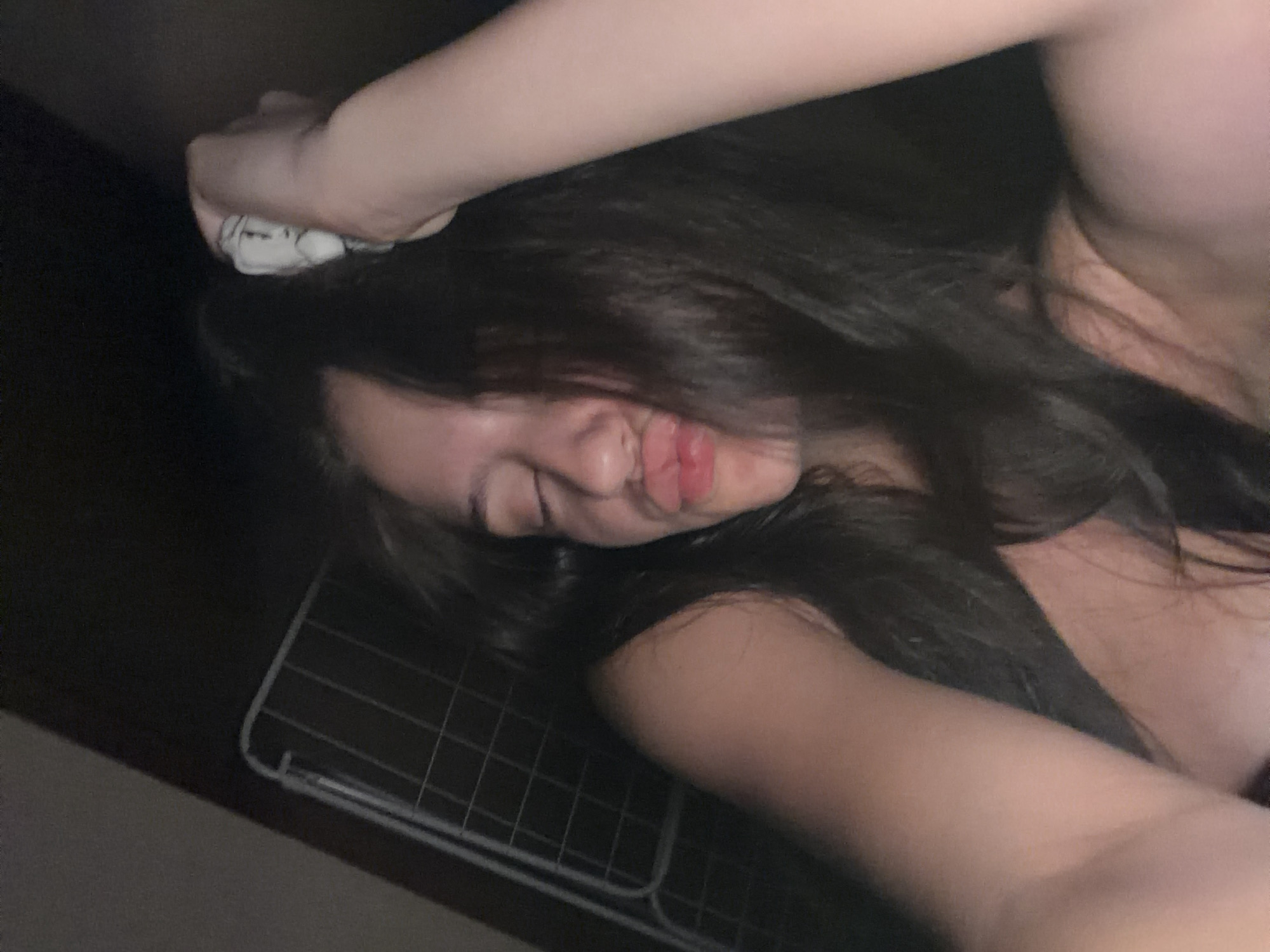 The image shows a close-up, low-light, candid shot of a young person with long, dark hair. Their eyes are closed, and their lips are pursed into a kiss-like expression. One arm is raised, holding a white scrunchie or hair accessory. The person appears to be leaning back, with their shoulder and upper chest visible. In the background, a metal grate or fence can be seen, suggesting an indoor or possibly enclosed outdoor space. The overall impression is of a casual, possibly playful moment captured at an intimate angle. There is no specific information to suggest a location within the city of Burgas, Bulgaria.