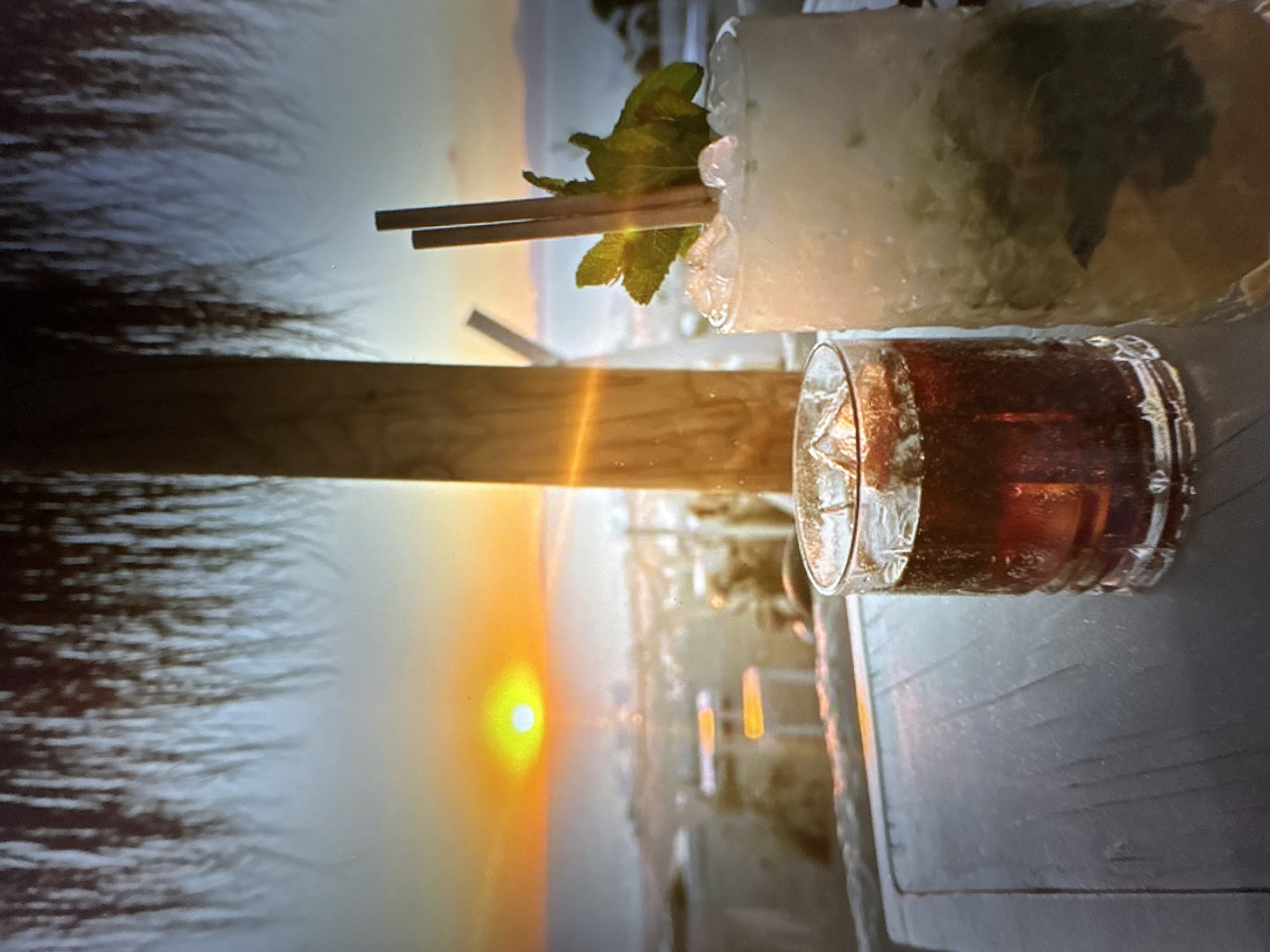 The image depicts two cocktails on a wooden table, with a blurred beach scene and sunset in the background. The left cocktail is a dark, carbonated beverage in a fluted glass filled with ice cubes. The right cocktail is a lighter-colored drink, also in a glass filled with crushed ice and garnished with mint leaves and two wooden stirrers. The background shows a sandy beach with lounge chairs and umbrellas, and a vibrant orange sun setting over the ocean. The scene is framed by what appears to be a thatched roof structure, suggesting an outdoor seating area at a beach bar or restaurant. There are no visible people. The location is not identifiable as Pori, Finland, as there are no distinguishing landmarks or features.