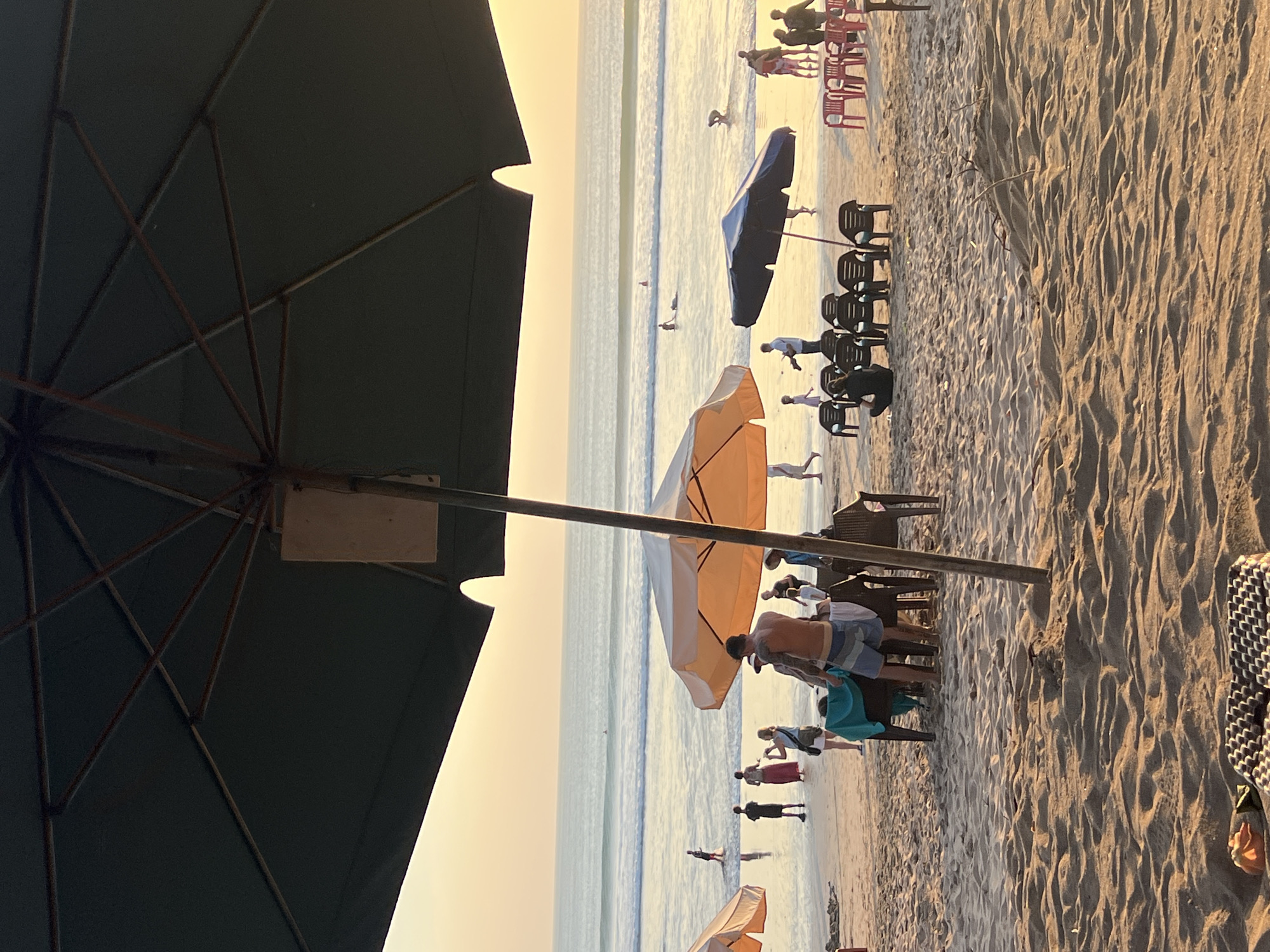 The image depicts a beach scene in Kuta, Indonesia, during what appears to be sunset. The foreground is dominated by sand with rippled textures from the wind and tide. Two large, dark umbrellas are prominent, one in the upper left and another in the center right, casting shade. Beneath the center umbrella, several chairs are arranged around tables, suggesting a seating area for a beachside establishment.
In the midground, a group of people are visible on the beach. To the left, under a lighter-colored umbrella, a man in swim trunks stands near tables and chairs, with other individuals, including a woman with a camera, also present. Further back, closer to the water, several people are walking and standing. One person is in the ocean, appearing to be surfing.
The background features the ocean with gentle waves rolling towards the shore. The sky is a soft, warm gradient of orange and yellow, indicative of the setting sun. The overall atmosphere is relaxed and leisurely, typical of a popular tourist beach.