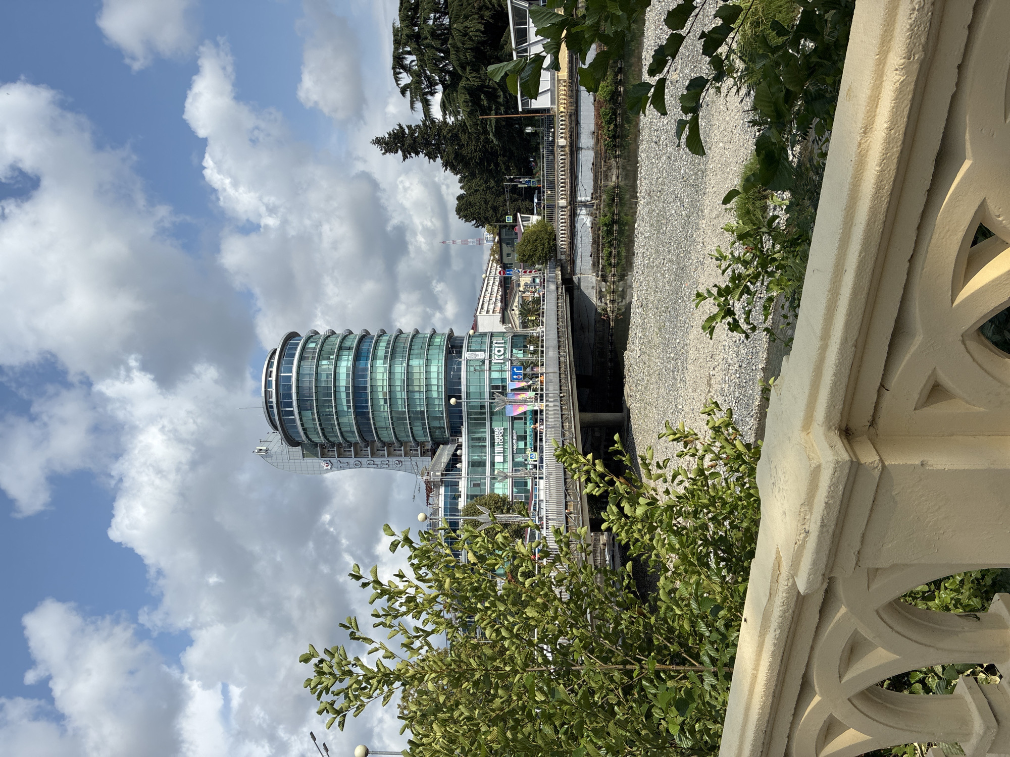 This image depicts a view of the city of Sochi, Russia, with a focus on the Novy Gorod district. In the foreground, a decorative beige stone railing with arched openings overlooks a riverbed filled with pebbles. Lush green foliage from trees and bushes partially obscures the view.
Dominating the midground is the distinctive, circular "Novy Gorod" building, characterized by its glass facade and modern architectural design. A bridge spans the river, with vehicles and pedestrians visible on it. The river itself appears shallow, with exposed pebble banks, suggesting a low water level.
In the background, more trees, buildings, and a telecommunications tower can be seen under a partly cloudy sky with scattered white clouds against a blue sky. The overall scene suggests a bright, clear day in an urban environment with a prominent river running through it. The "Novy Gorod" building is a key landmark in this part of Sochi.